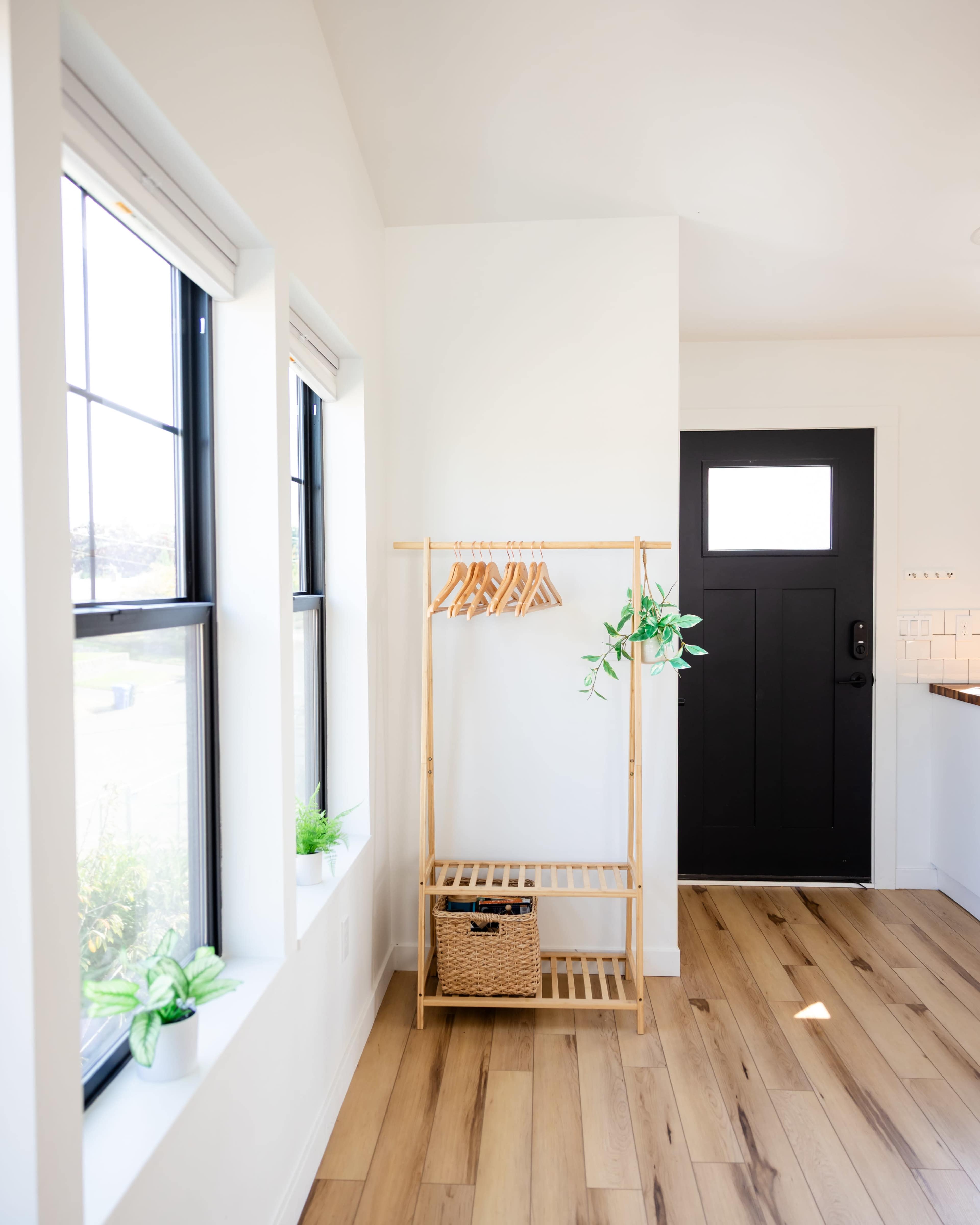 The image depicts a modern entryway featuring a wooden coat rack, a storage basket, and two windows alongside a black front door.