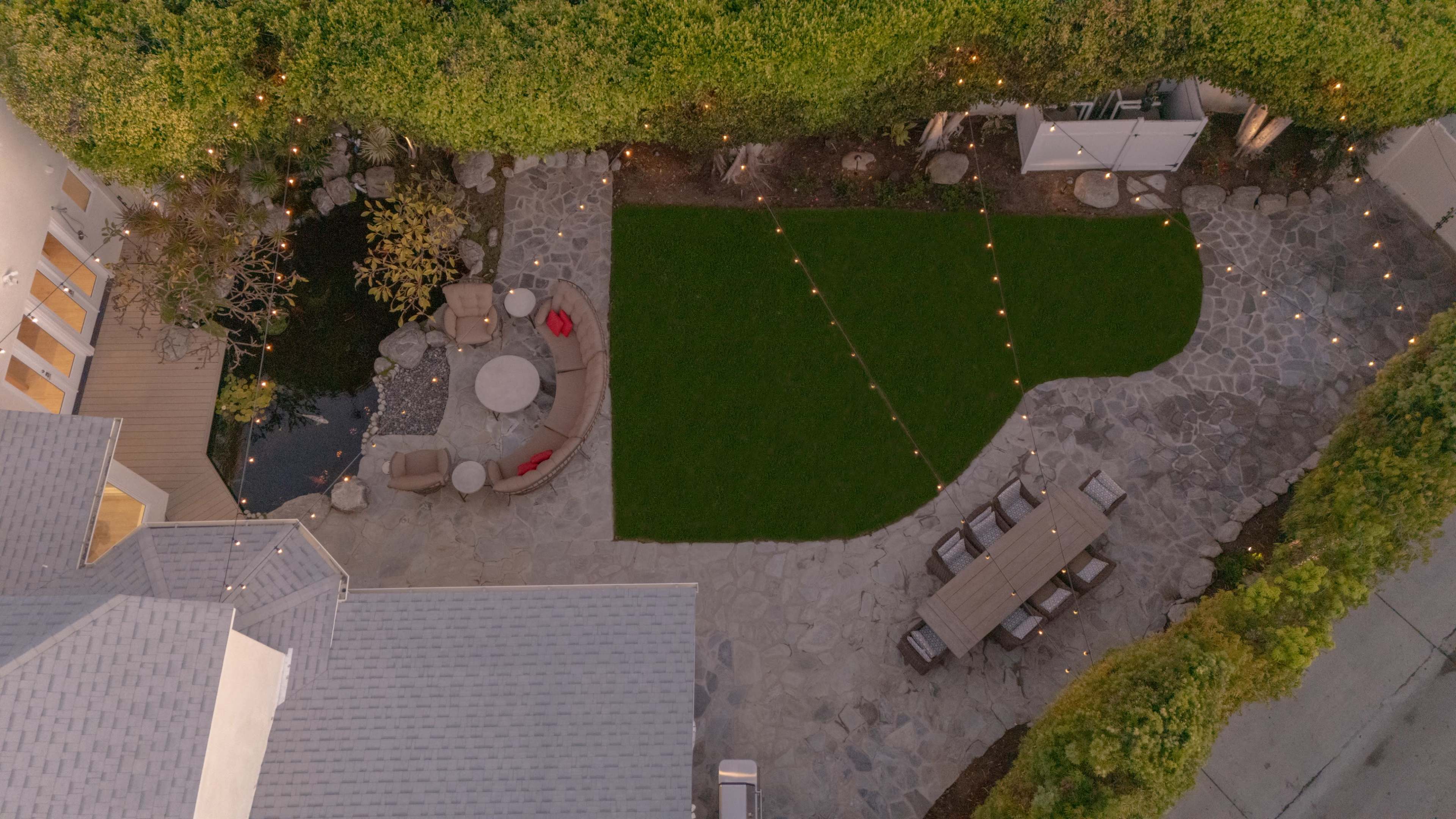 The image shows an aerial view of a landscaped backyard with a seating area, a stone patio, a lawn, and string lights overhead.