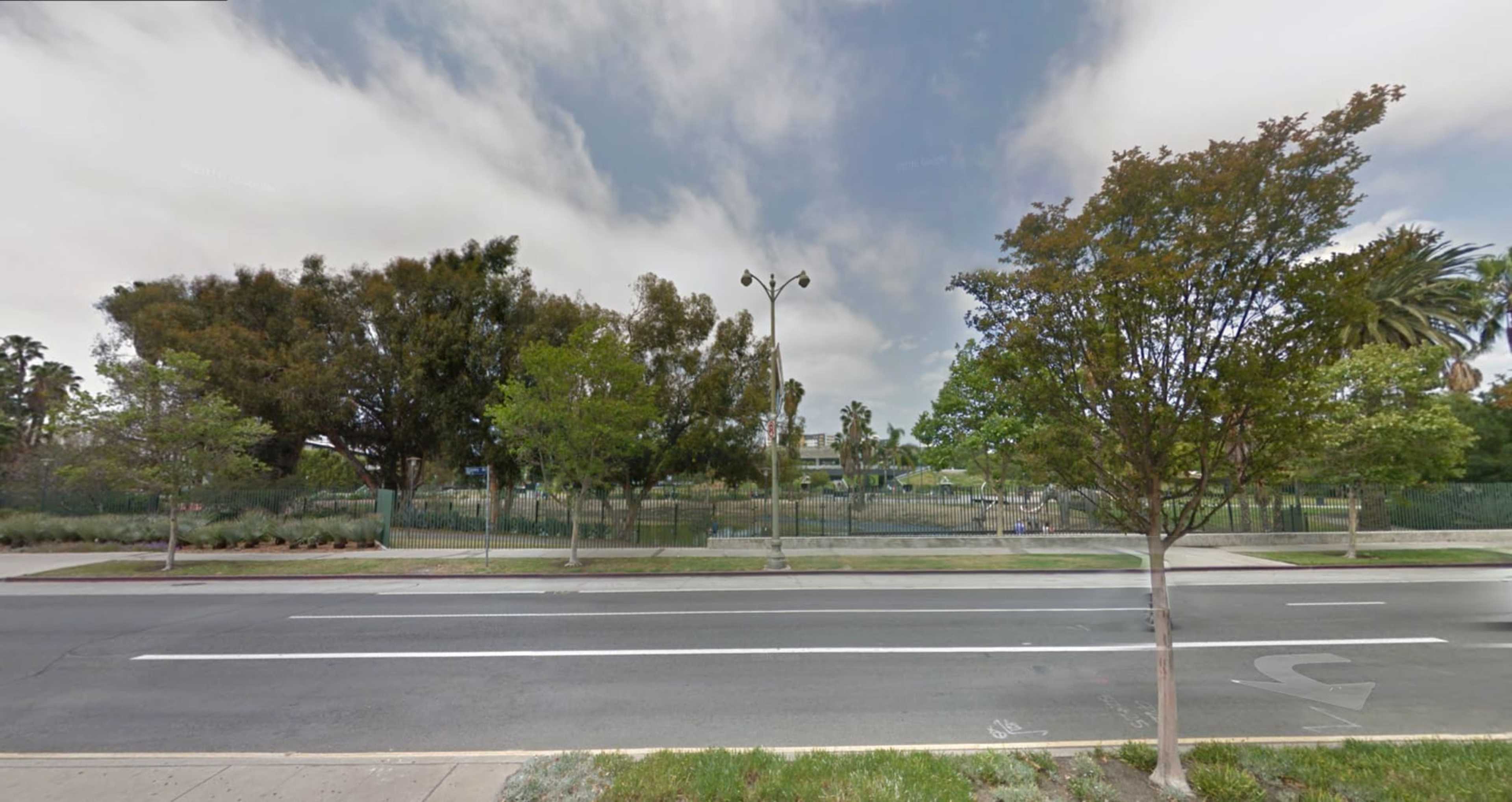 A street view with a line of trees and a fenced area in the background, under a cloudy sky.