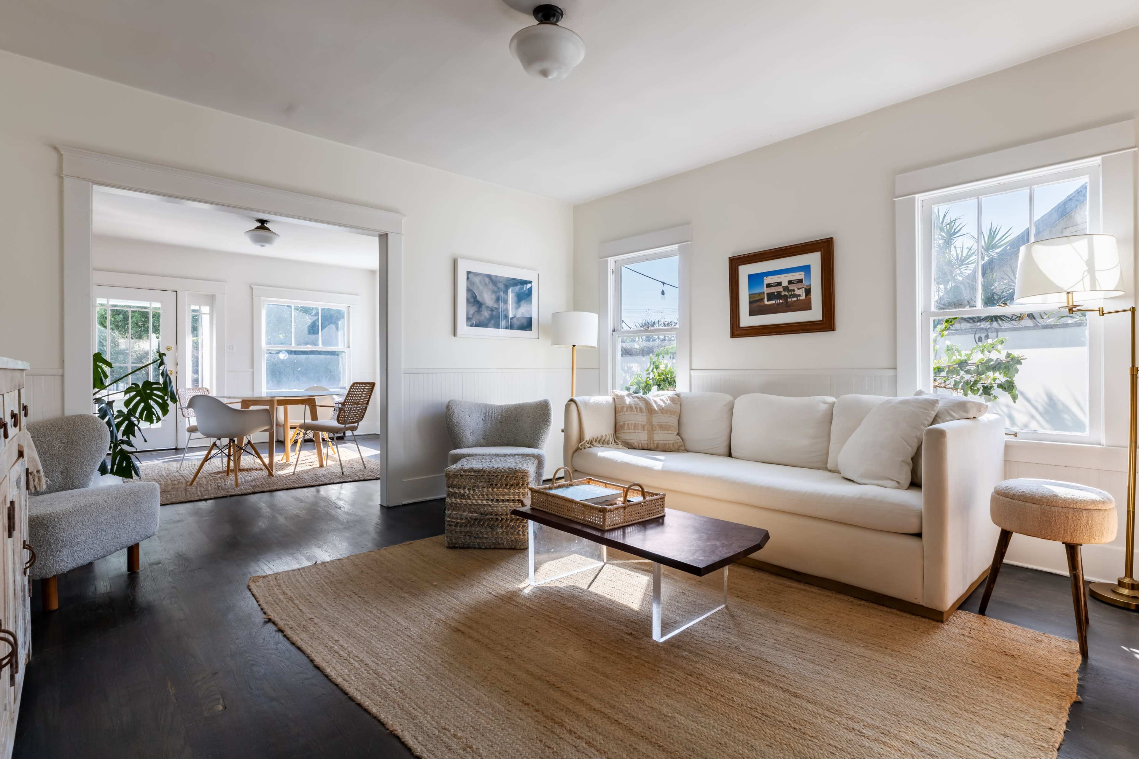 A bright living room features a beige sofa, a wooden coffee table, and large windows that let in natural light, with an adjacent dining area visible through an open doorway.