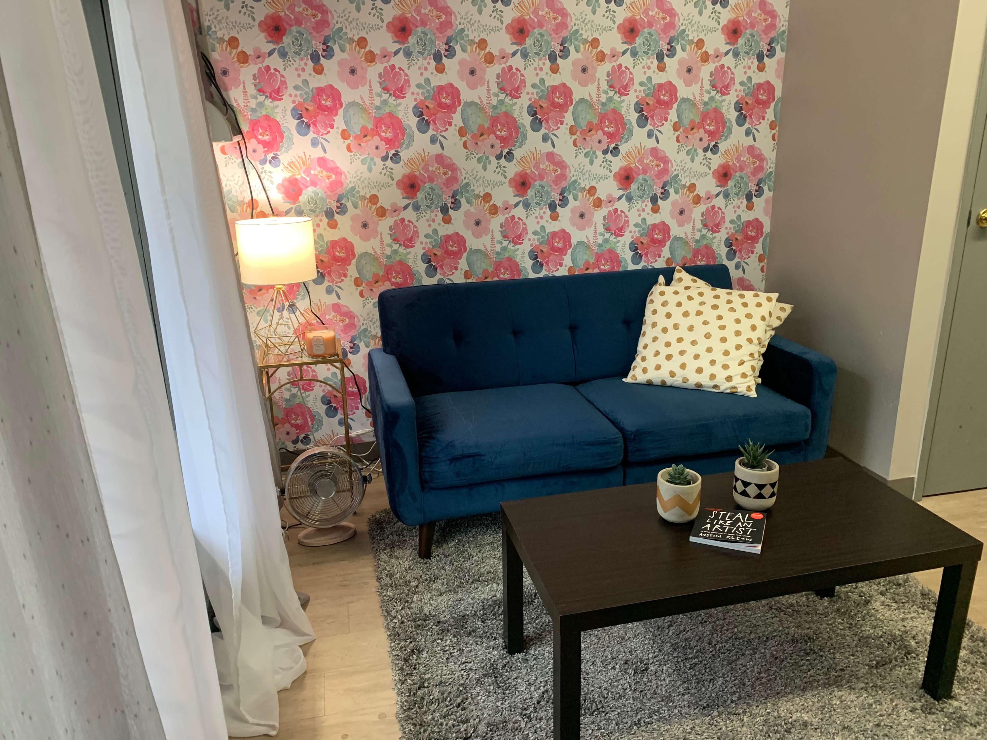 A living room with a blue sofa, a patterned wall, a coffee table, a lamp, and a small fan.