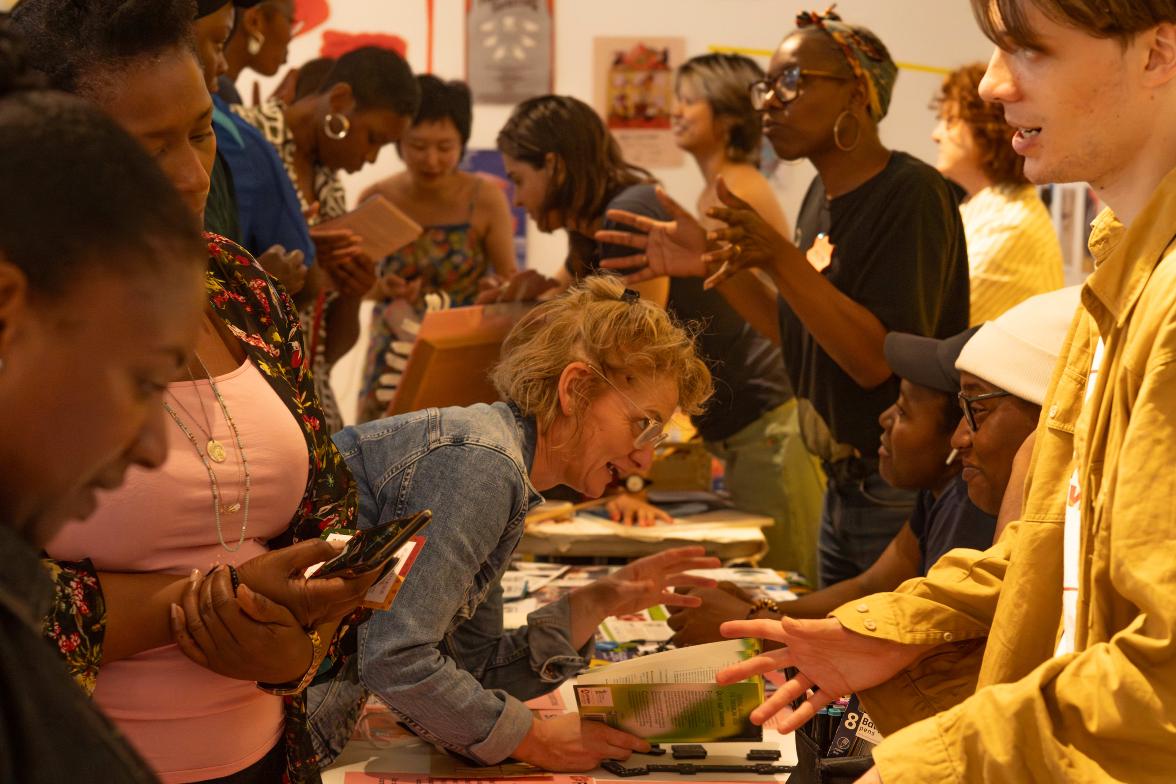 A diverse group of people engages in conversation and exchanges materials at tables in a vibrant indoor space filled with artwork and publications.