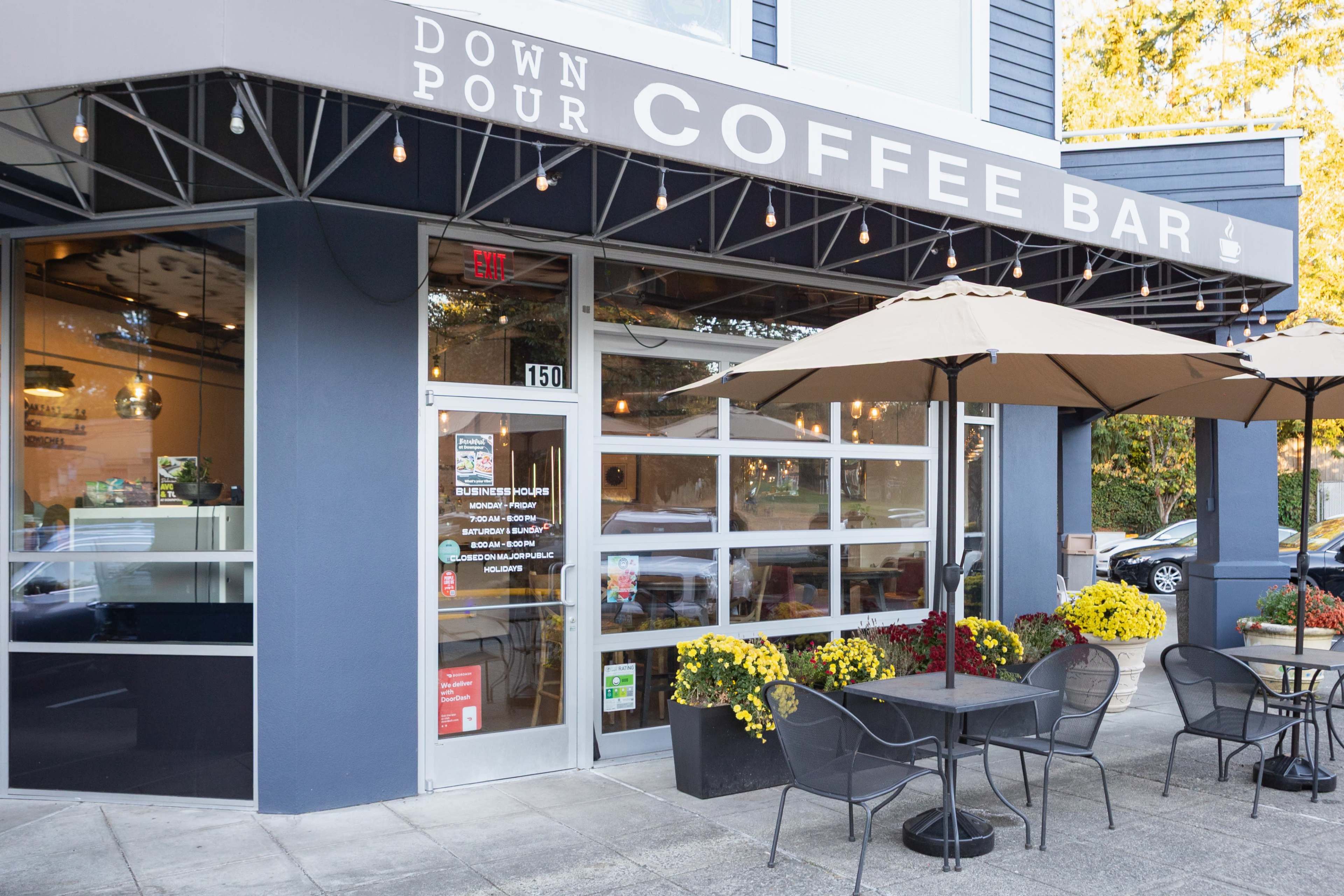 The image shows a coffee bar named "Down Pour Coffee Bar," featuring outdoor seating with tables and umbrellas, and flower pots adorned with autumn blooms.