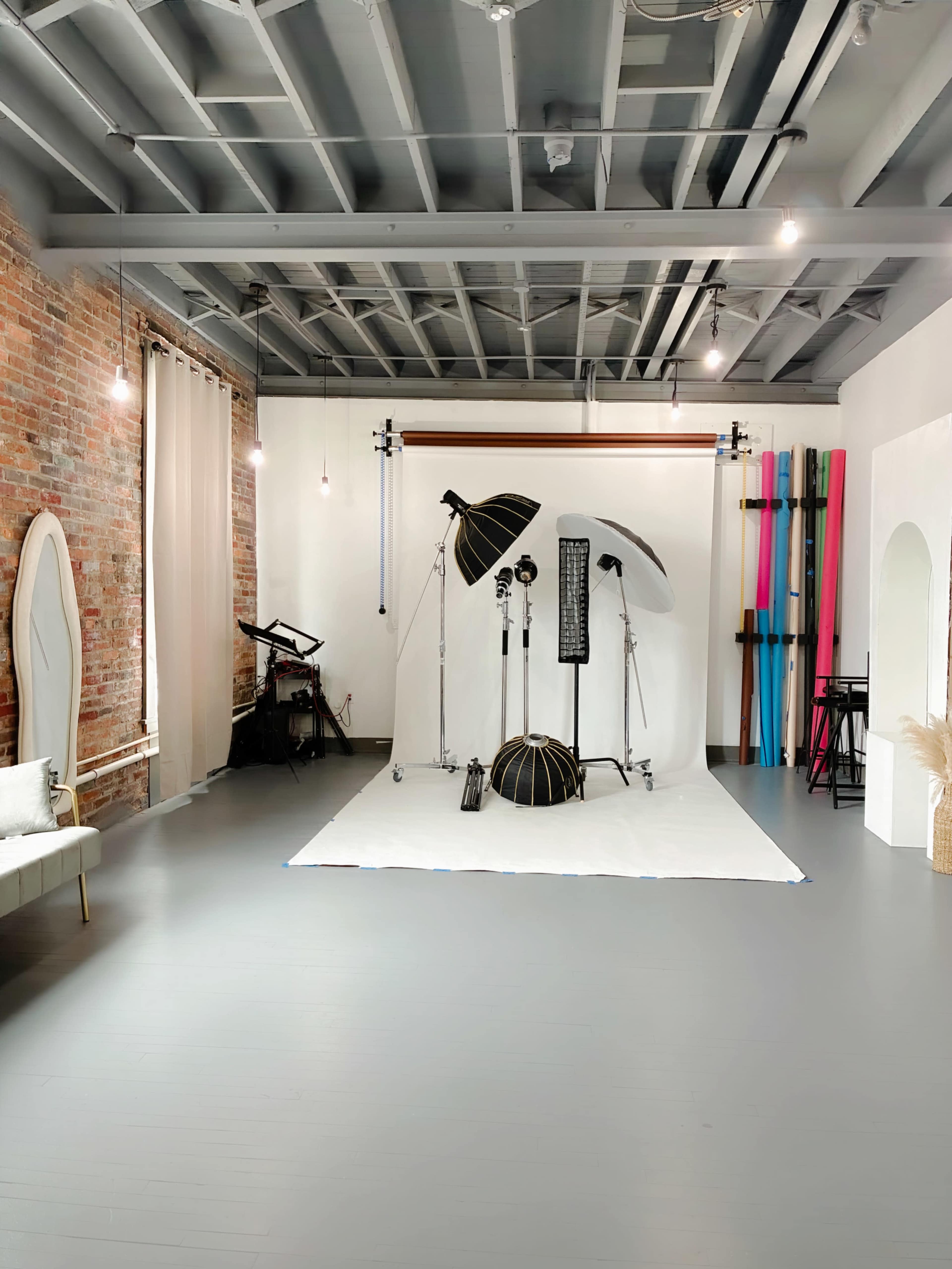 The image shows a photography studio with a white backdrop, studio lighting equipment, and colorful props arranged in a spacious, well-lit room with exposed brick walls.