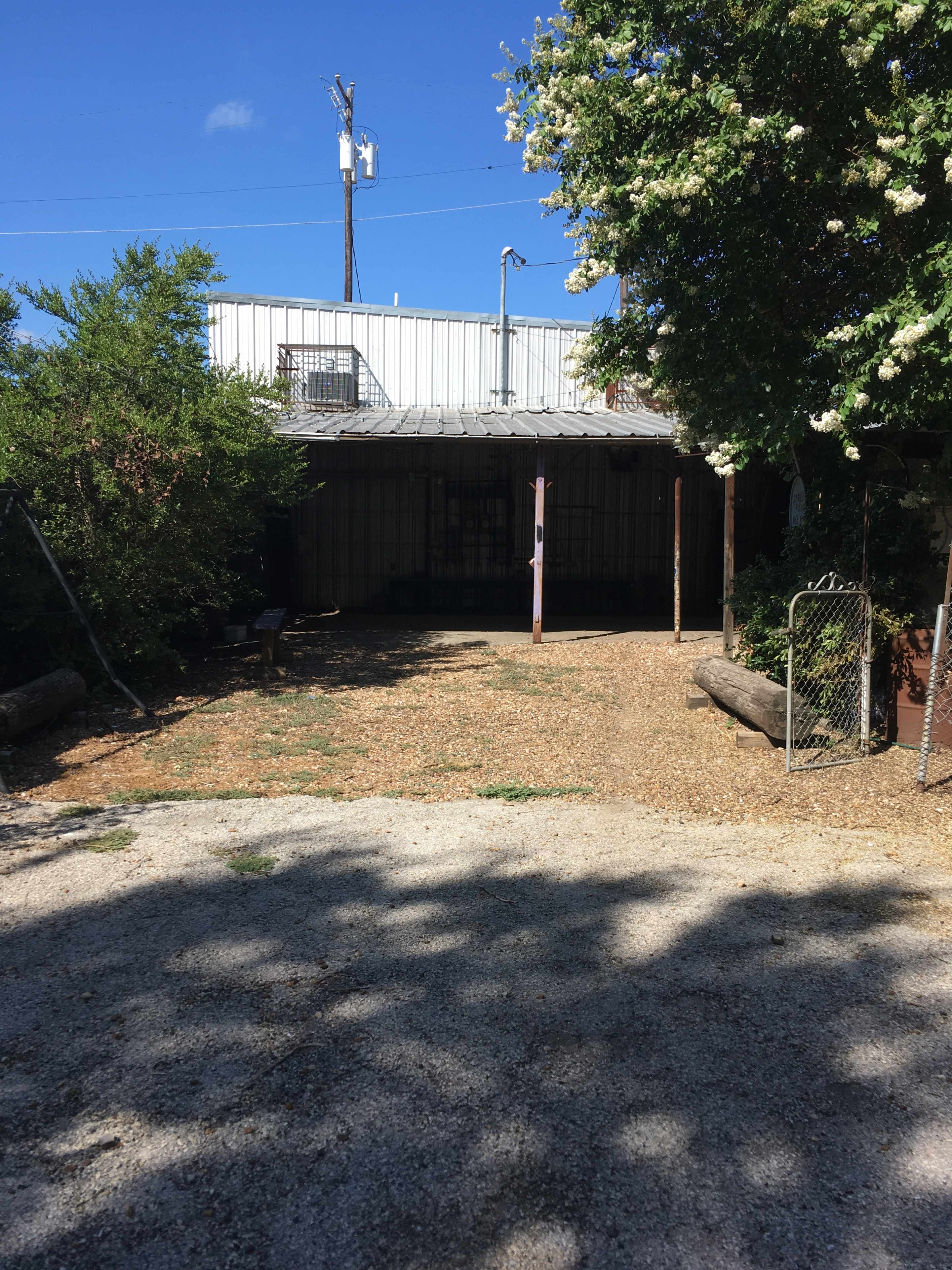 A gravel area in front of a metal building, partially shaded by trees, with a swing set on one side.