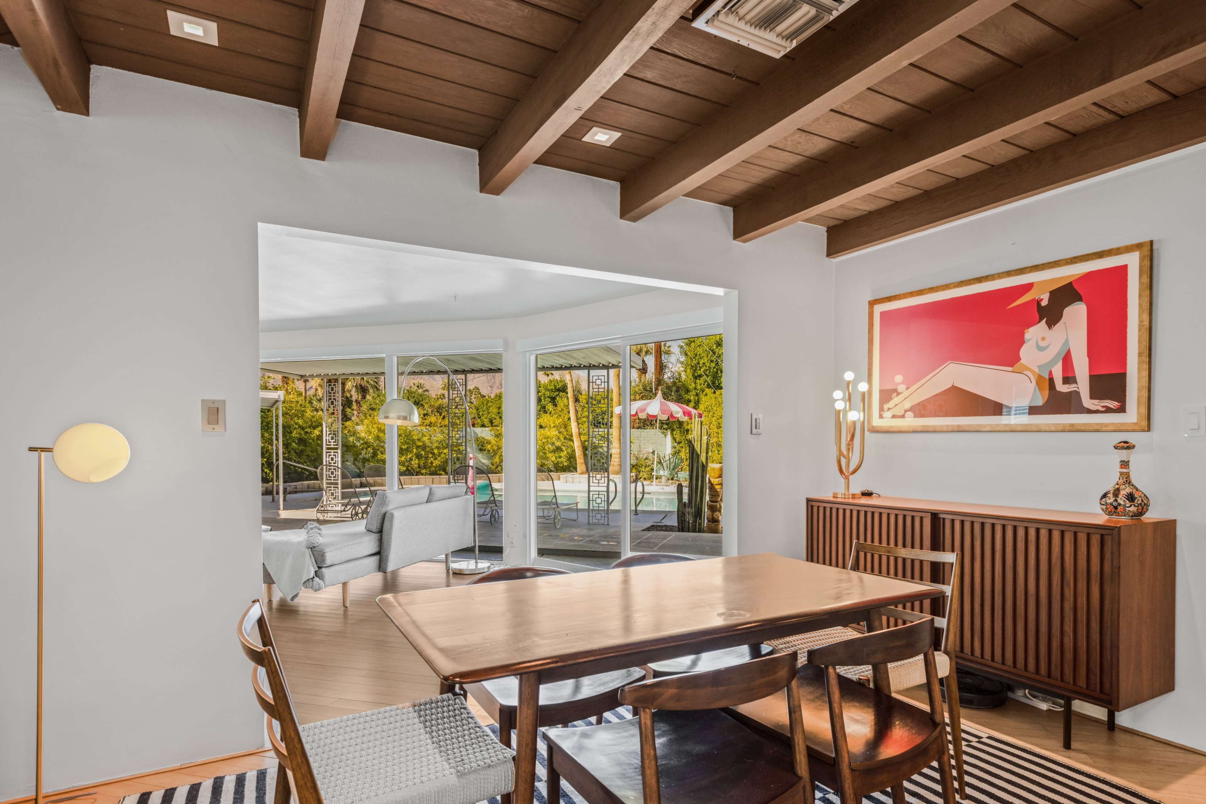 A dining area with a wooden table and chairs opens into a bright space featuring large windows and a view of a patio and pool.