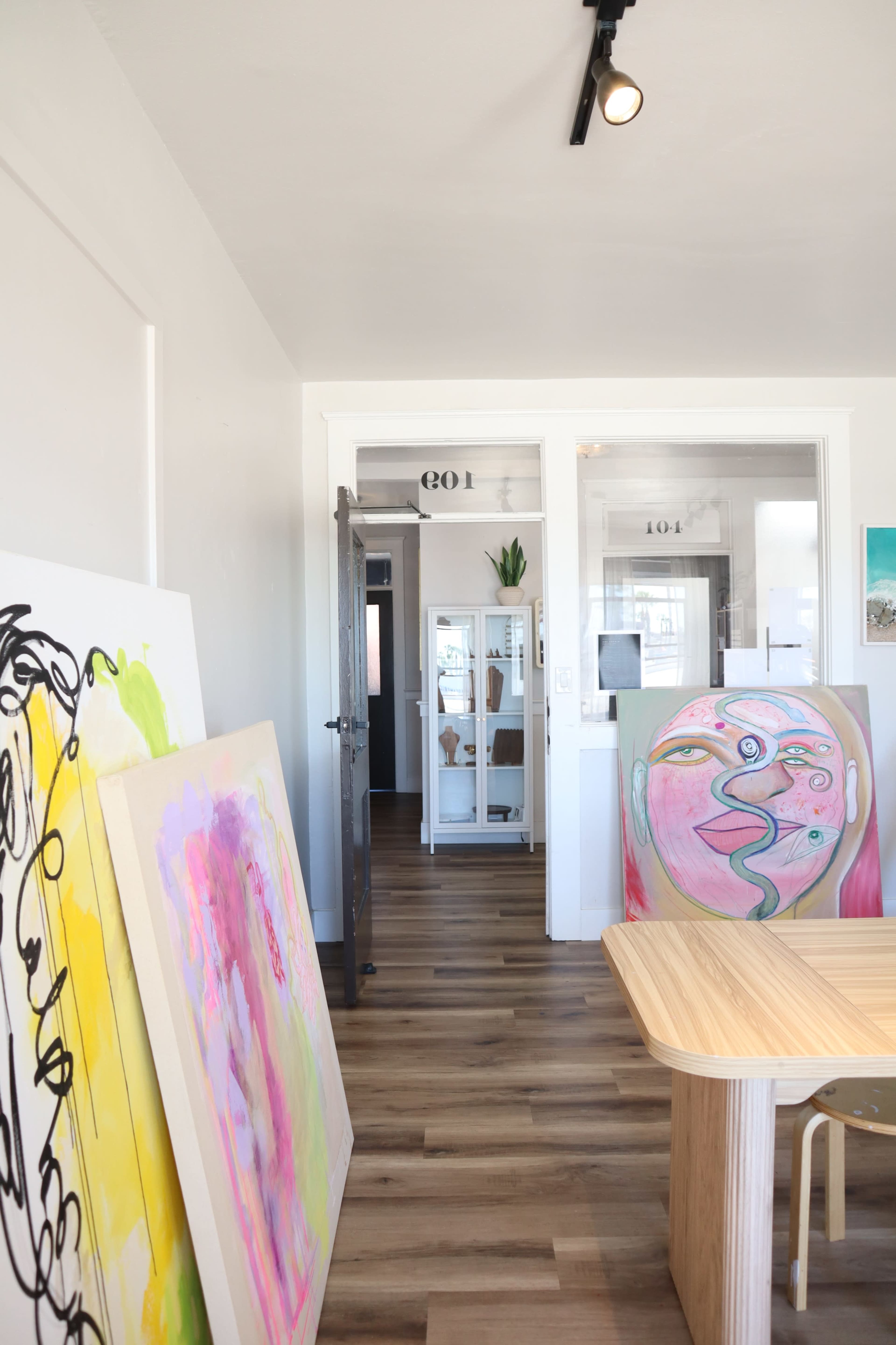 The image shows a spacious interior with colorful paintings against the wall and a wooden table in the foreground, leading to a bright room with glass door entries labeled "104" and "104."