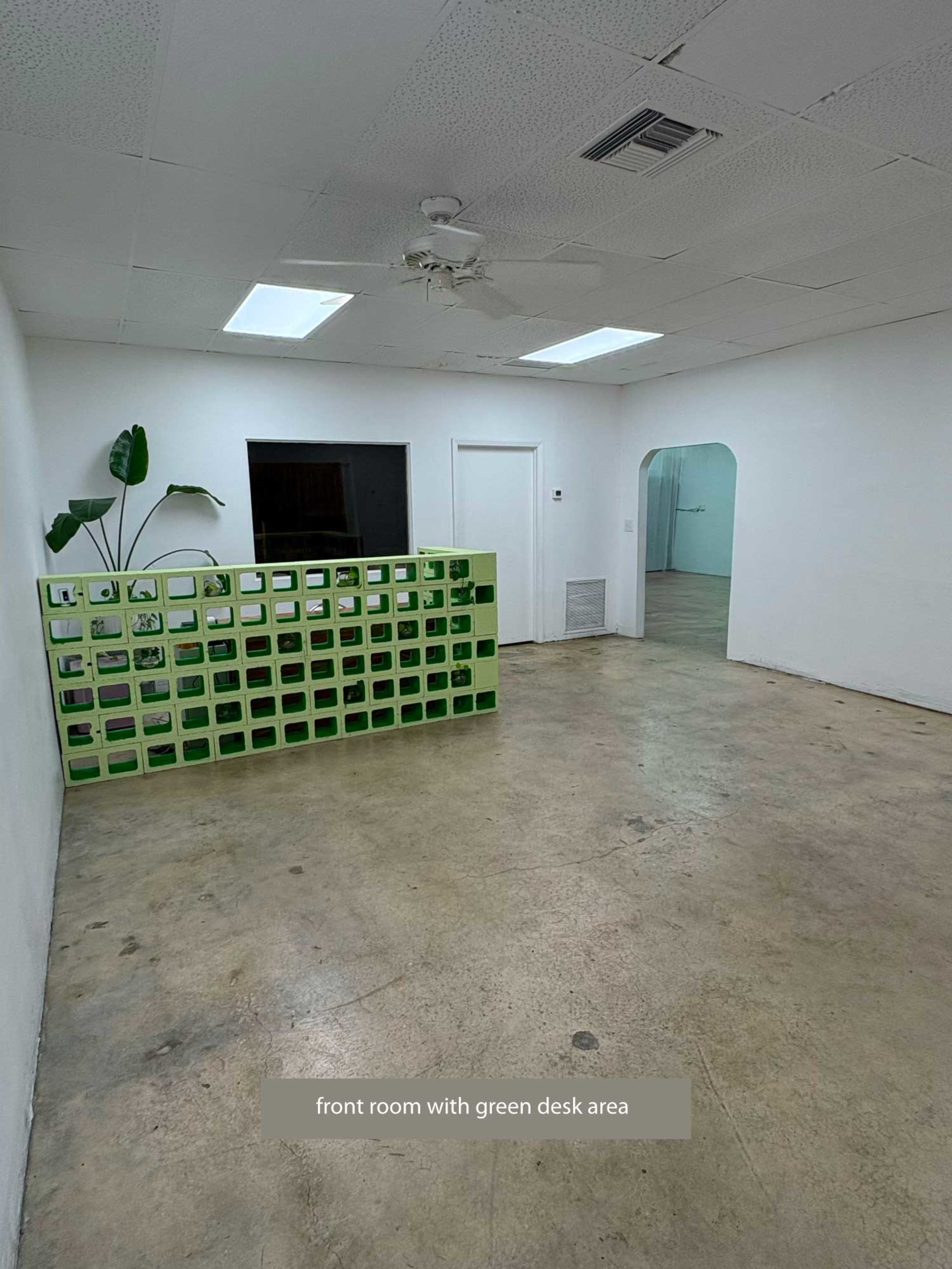 The image shows a front room with a green desk area defined by a low green partition, featuring white walls and a ceiling fan.