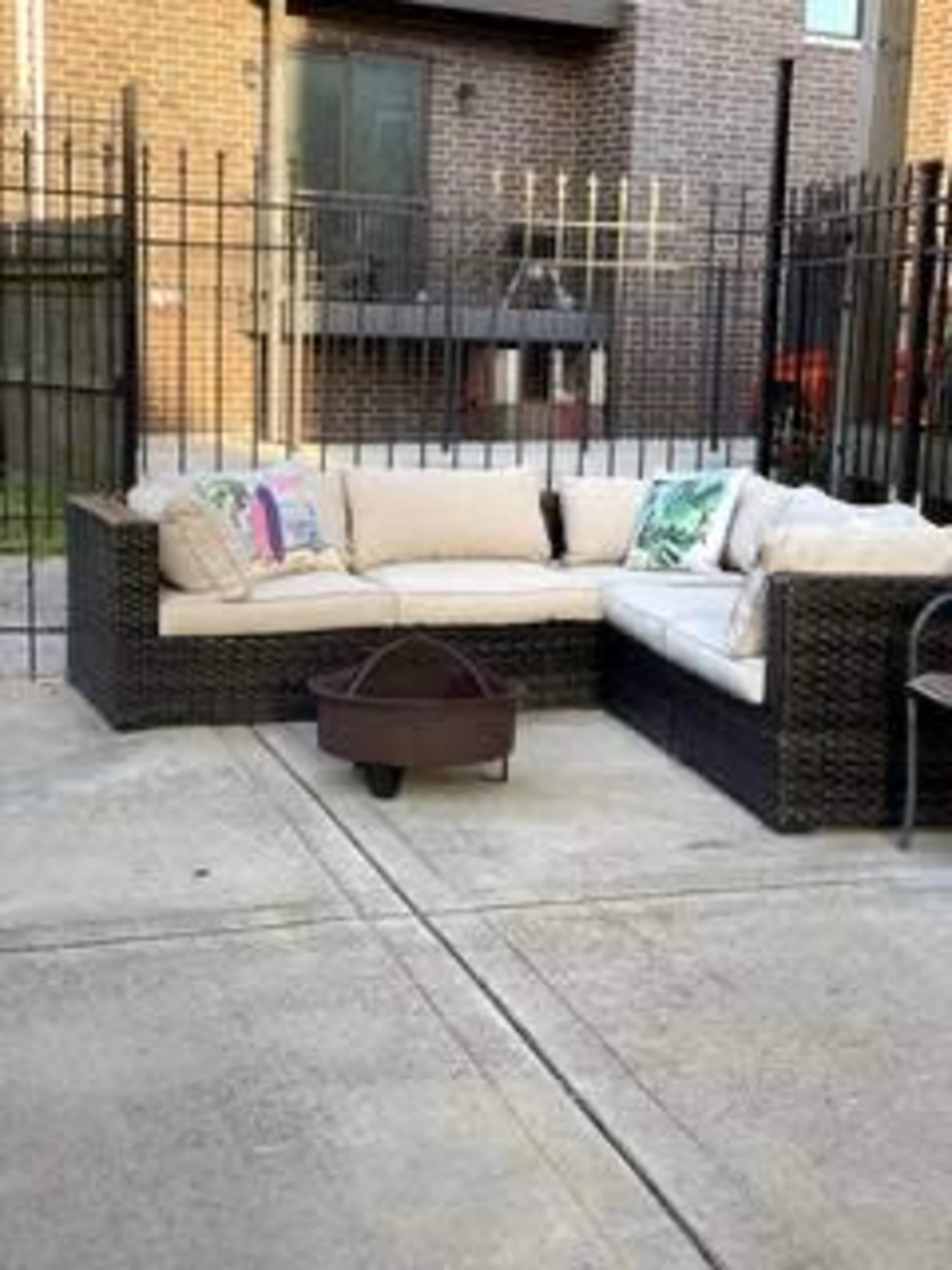 A wicker sectional sofa with cushions and a fire pit is set on a concrete patio behind a black metal fence.
