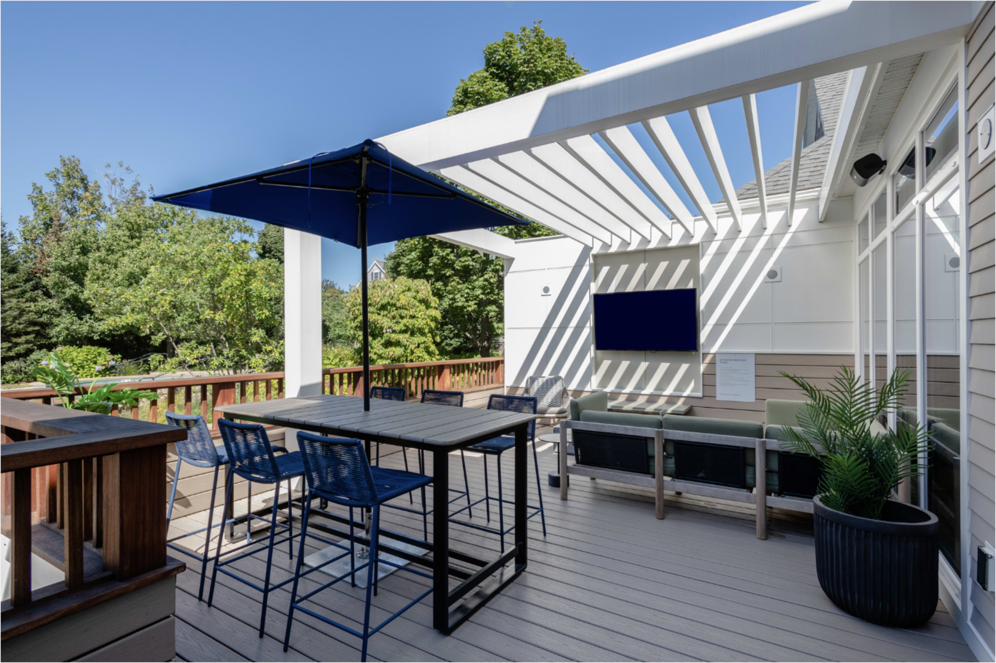 The image shows a modern outdoor patio with a shaded seating area, a table with high stools, a blue umbrella, and a television mounted on the wall.