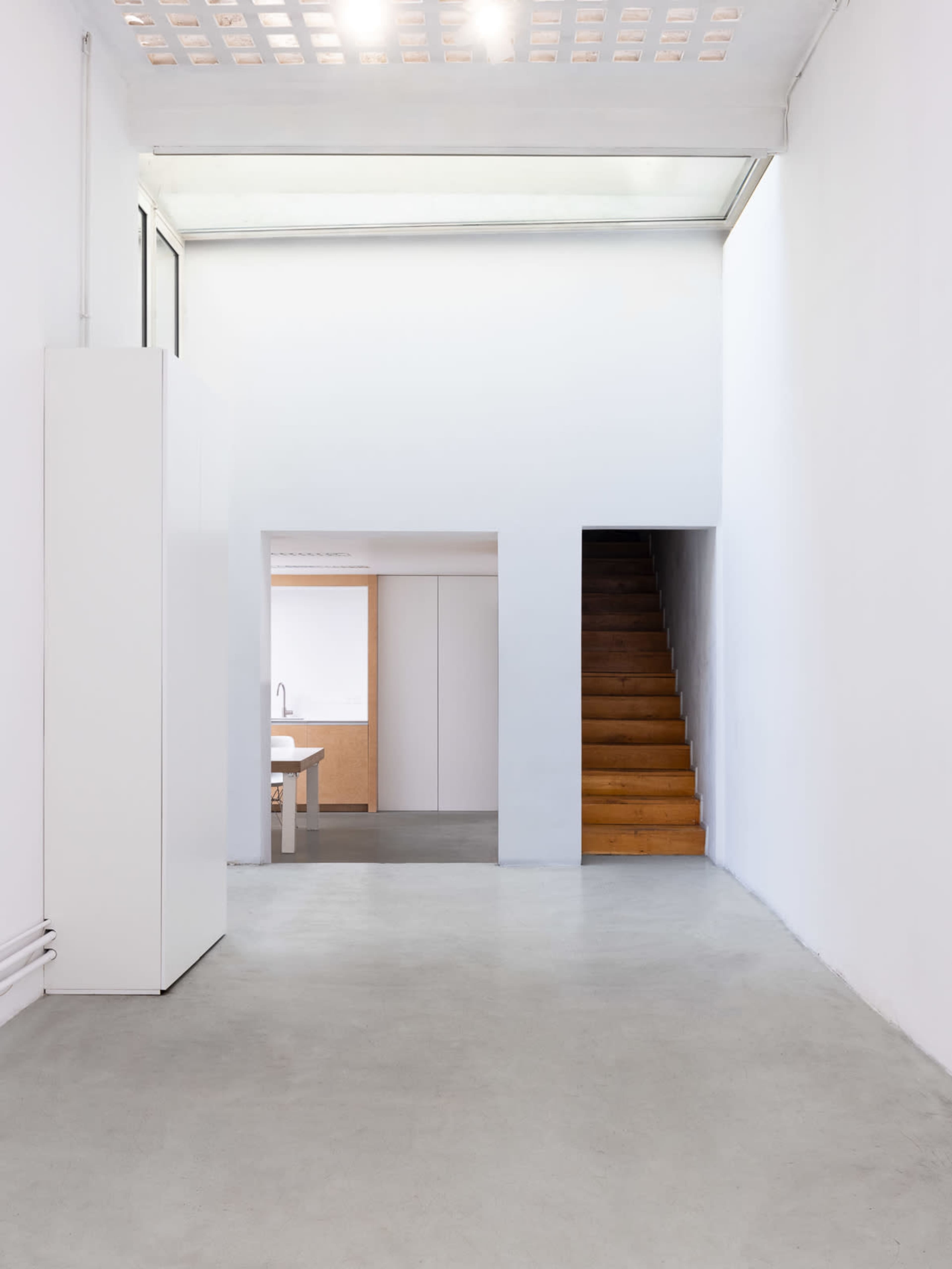 The image shows a minimalistic interior space with white walls, a wooden staircase, and a kitchen area partially visible in the background.