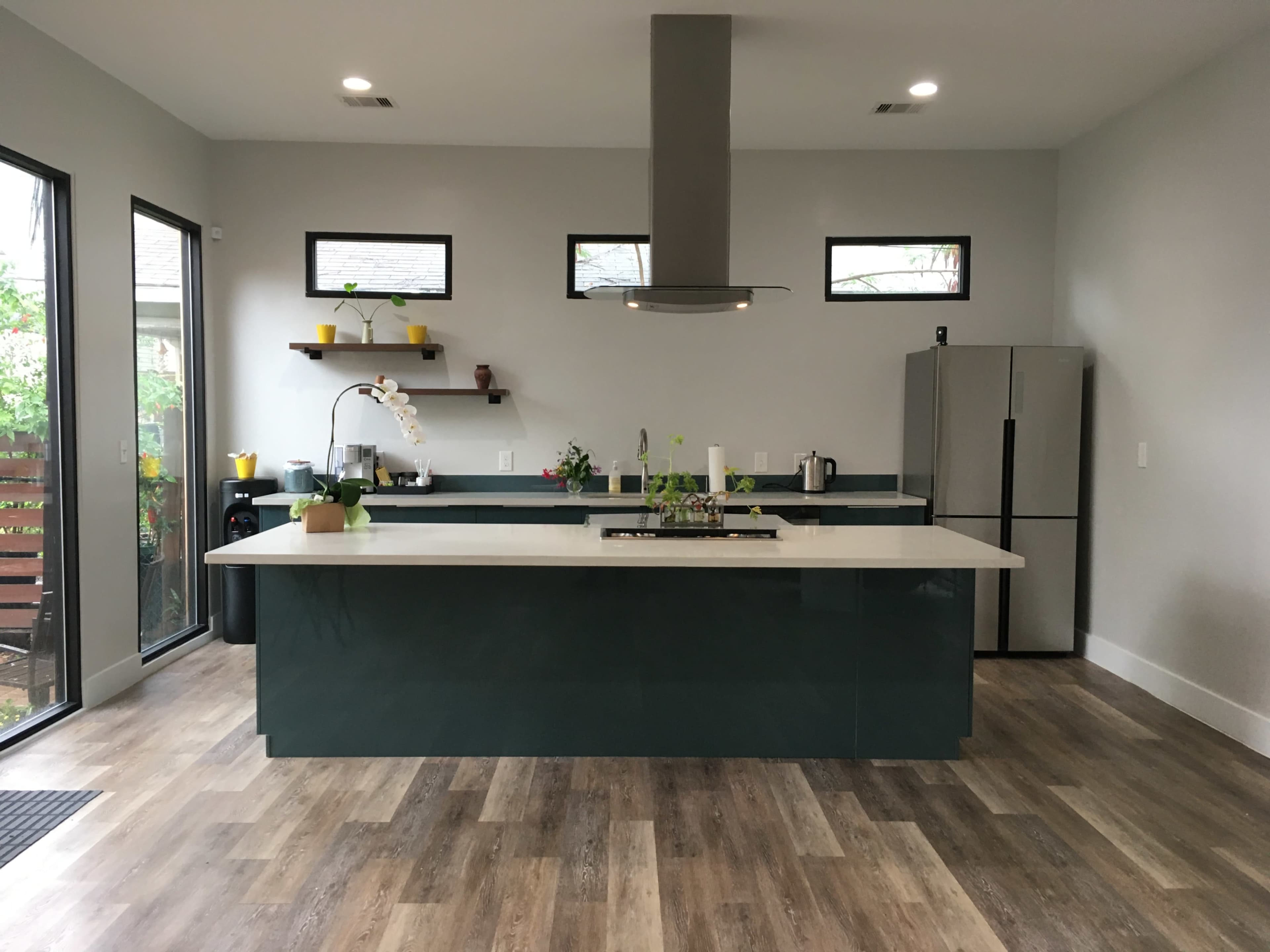A modern kitchen features a central island with a stovetop, a stainless steel refrigerator, and large windows allowing natural light.