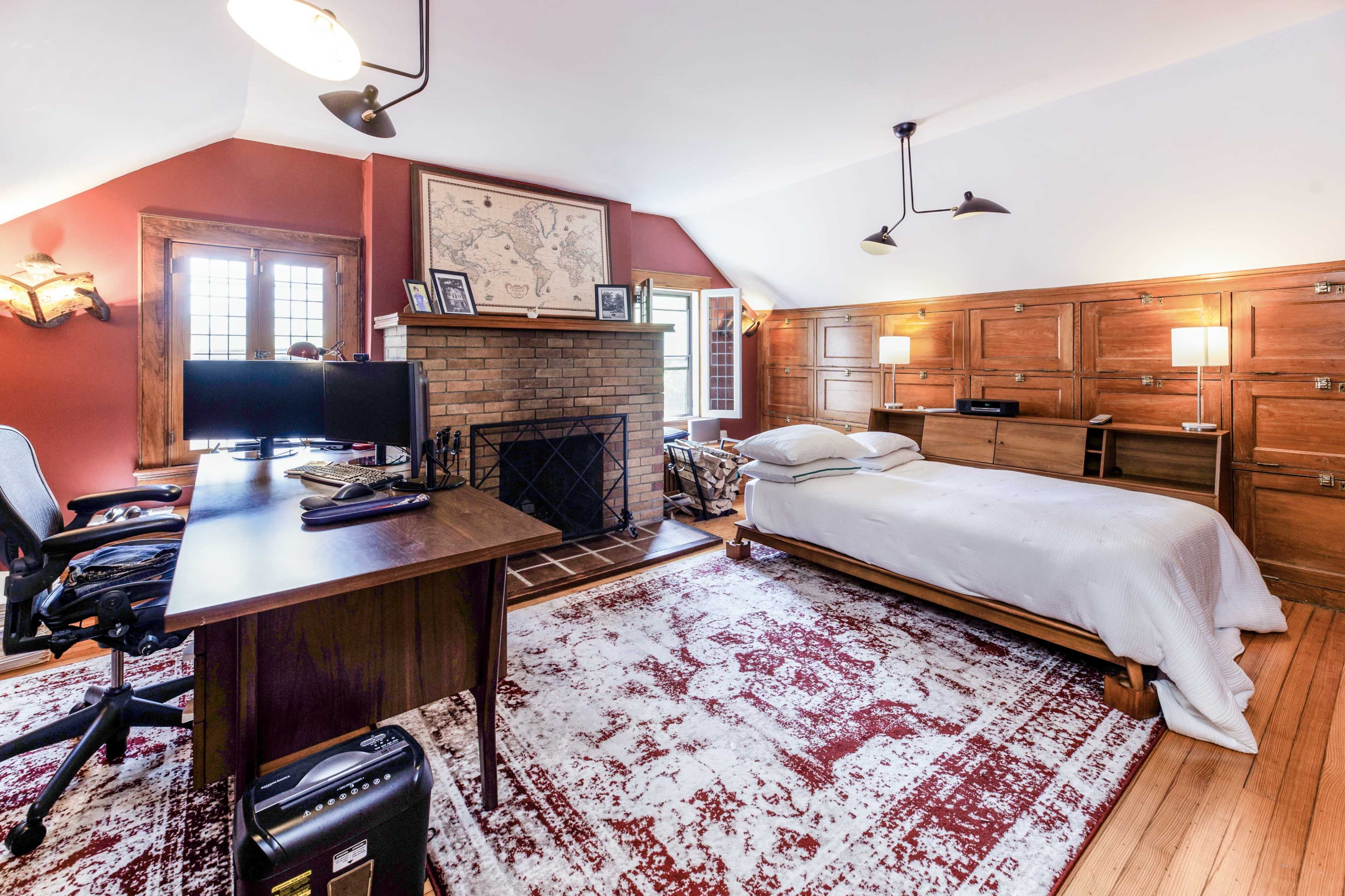 A bedroom with a workspace featuring two monitors, a bed, a fireplace with a map above it, and a patterned rug on the wooden floor.