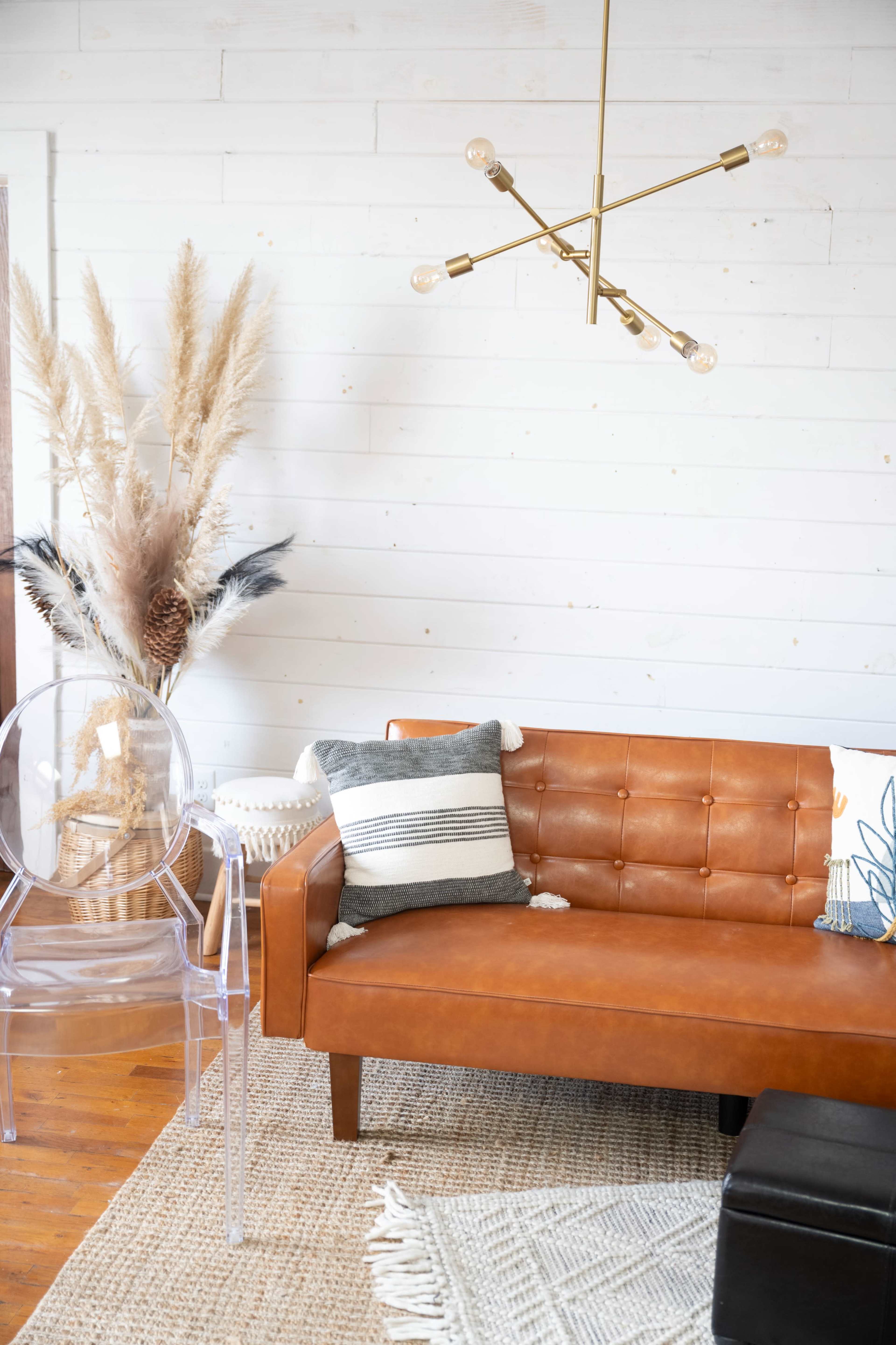 A brown leather sofa with tufted detailing sits next to a clear acrylic chair and decorative plants in a room with white walls.