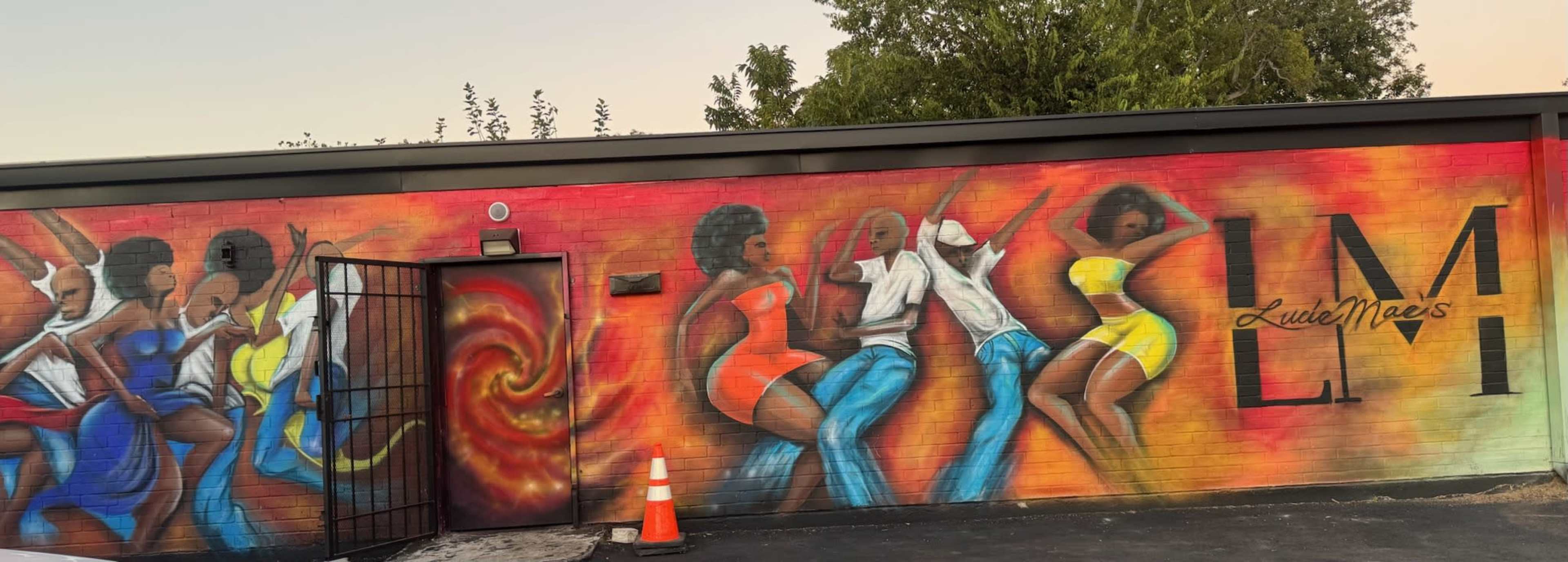 The image shows a colorful mural on a wall featuring dancing figures in vibrant attire alongside the text "Lucie Mae's."