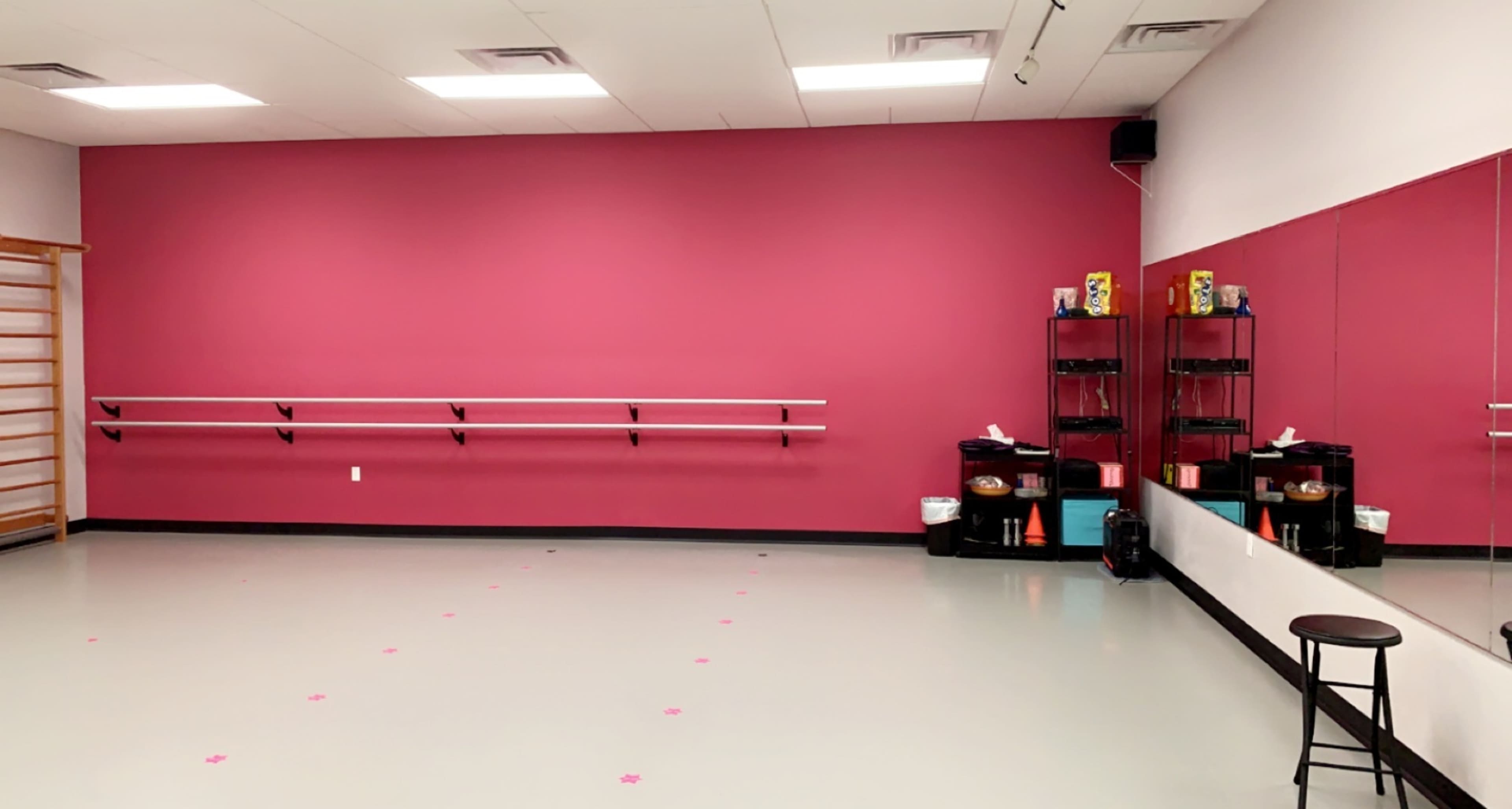 A dance studio with a pink wall, a mirrored section, a barre mounted on the wall, and a stool in one corner.