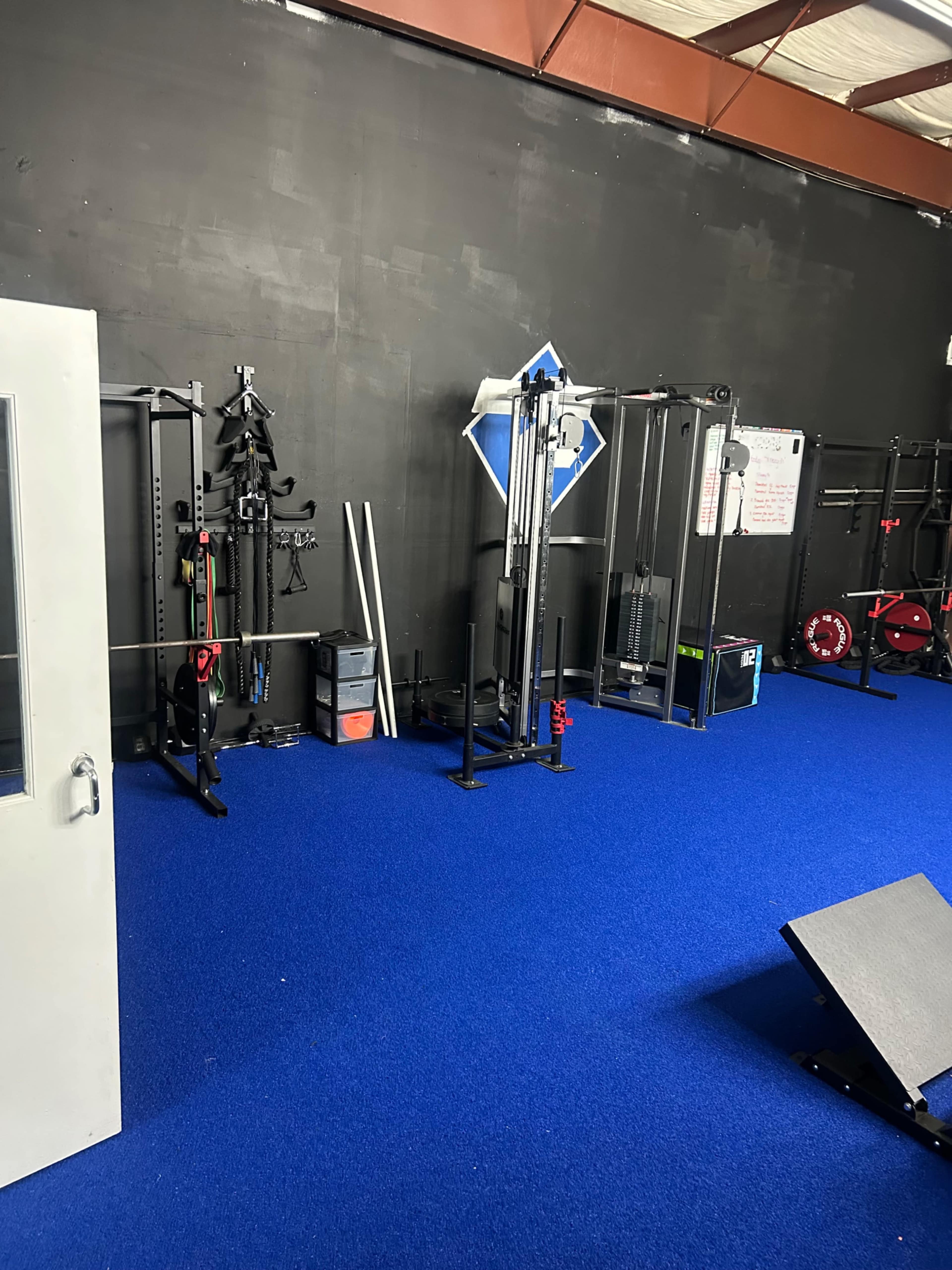 The image shows a gym interior featuring various workout equipment arranged against a black wall, with a blue carpeted floor.