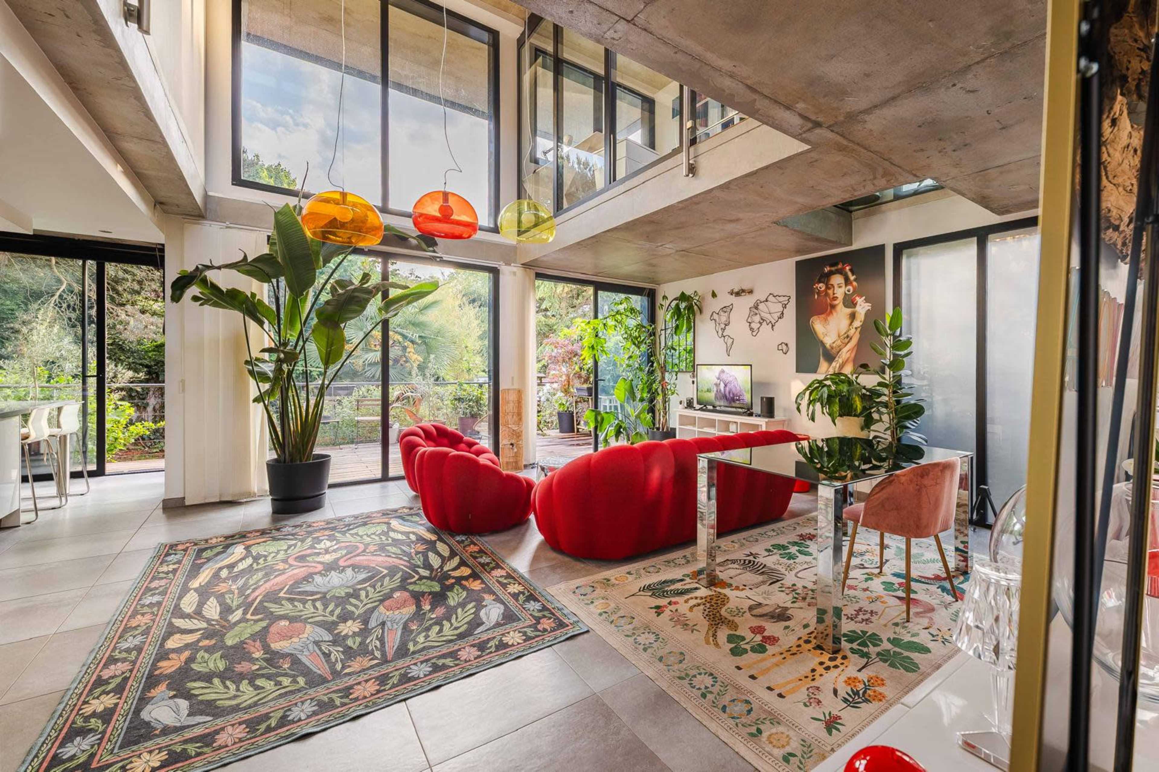 The image shows a modern living space with large windows, featuring red furniture, vibrant decor, and indoor plants.