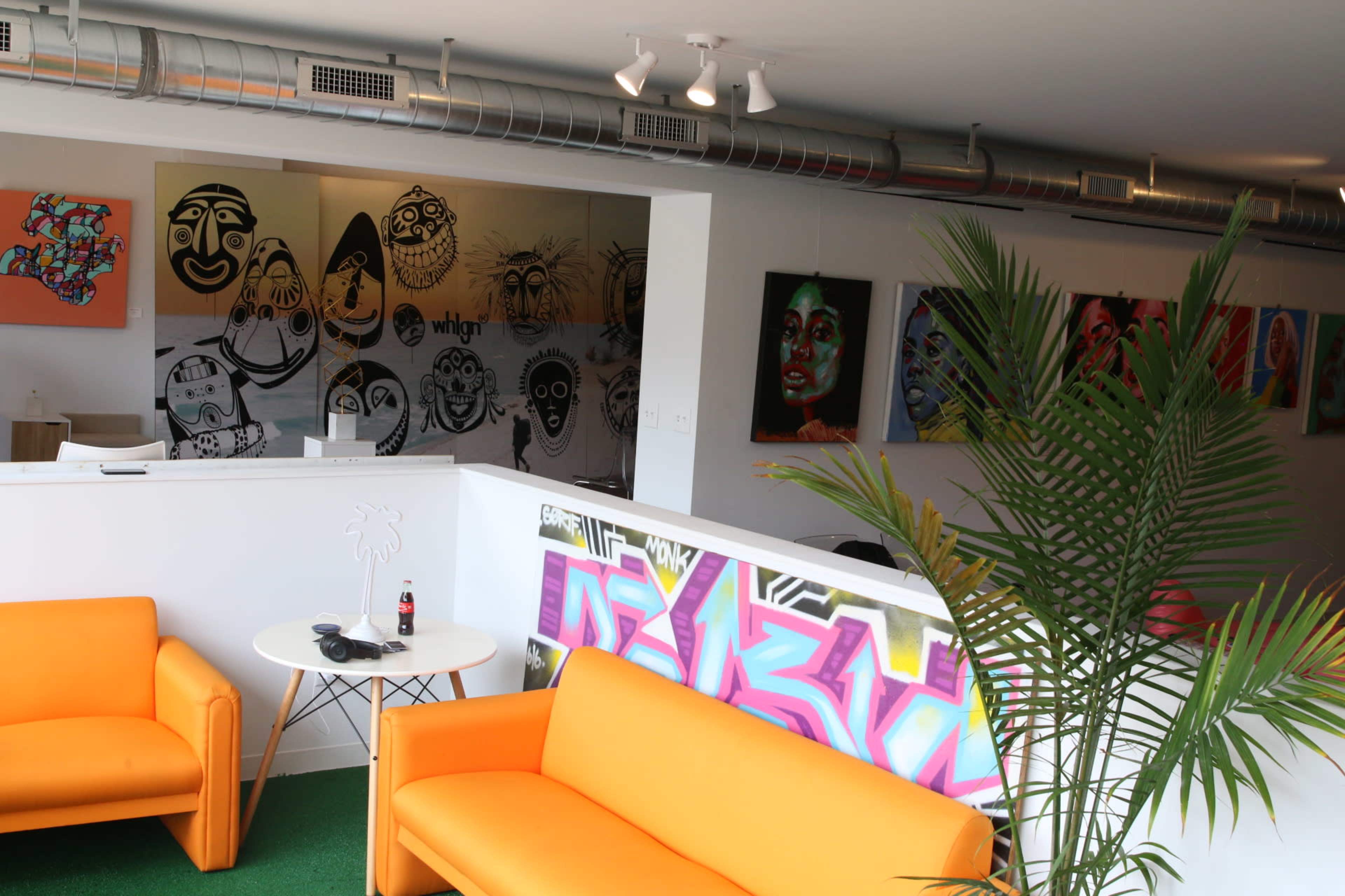 A modern office space with two orange sofas, a white partition featuring graffiti art, and several colorful paintings of faces on the walls.
