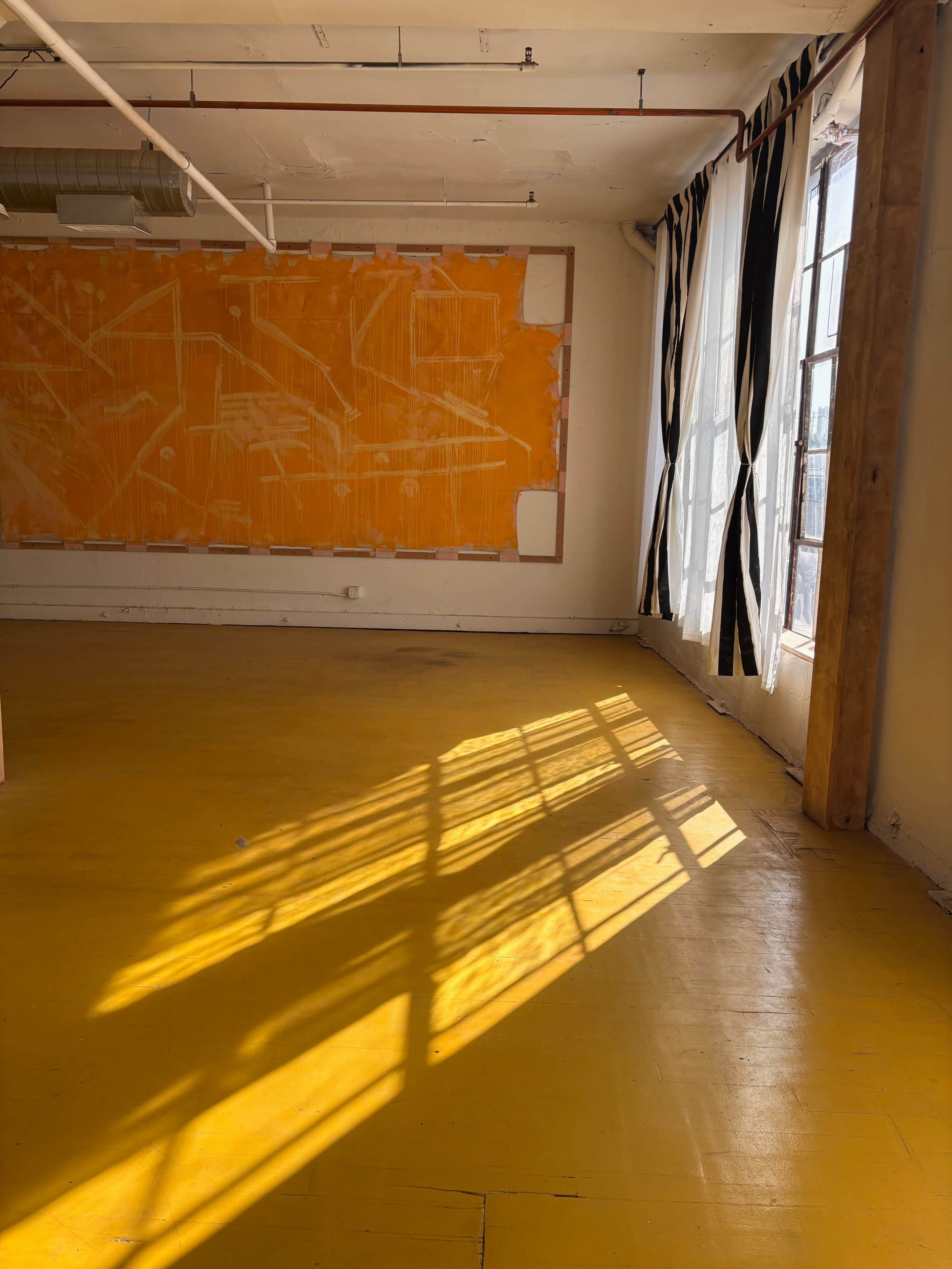 The image shows an empty room with yellow flooring, large windows casting shadows, and an orange wall in the background.