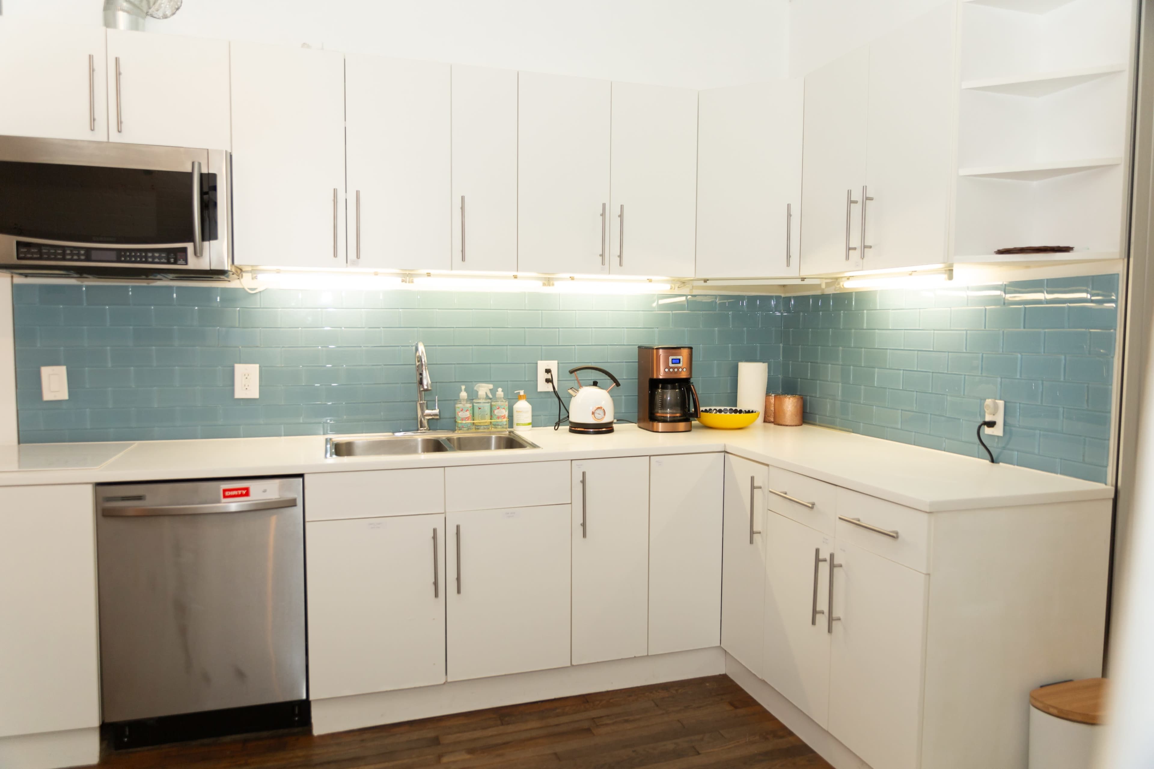 The image shows a modern kitchen with white cabinetry, a stainless steel dishwasher, a sink, and a coffee maker against a light blue tiled backsplash.