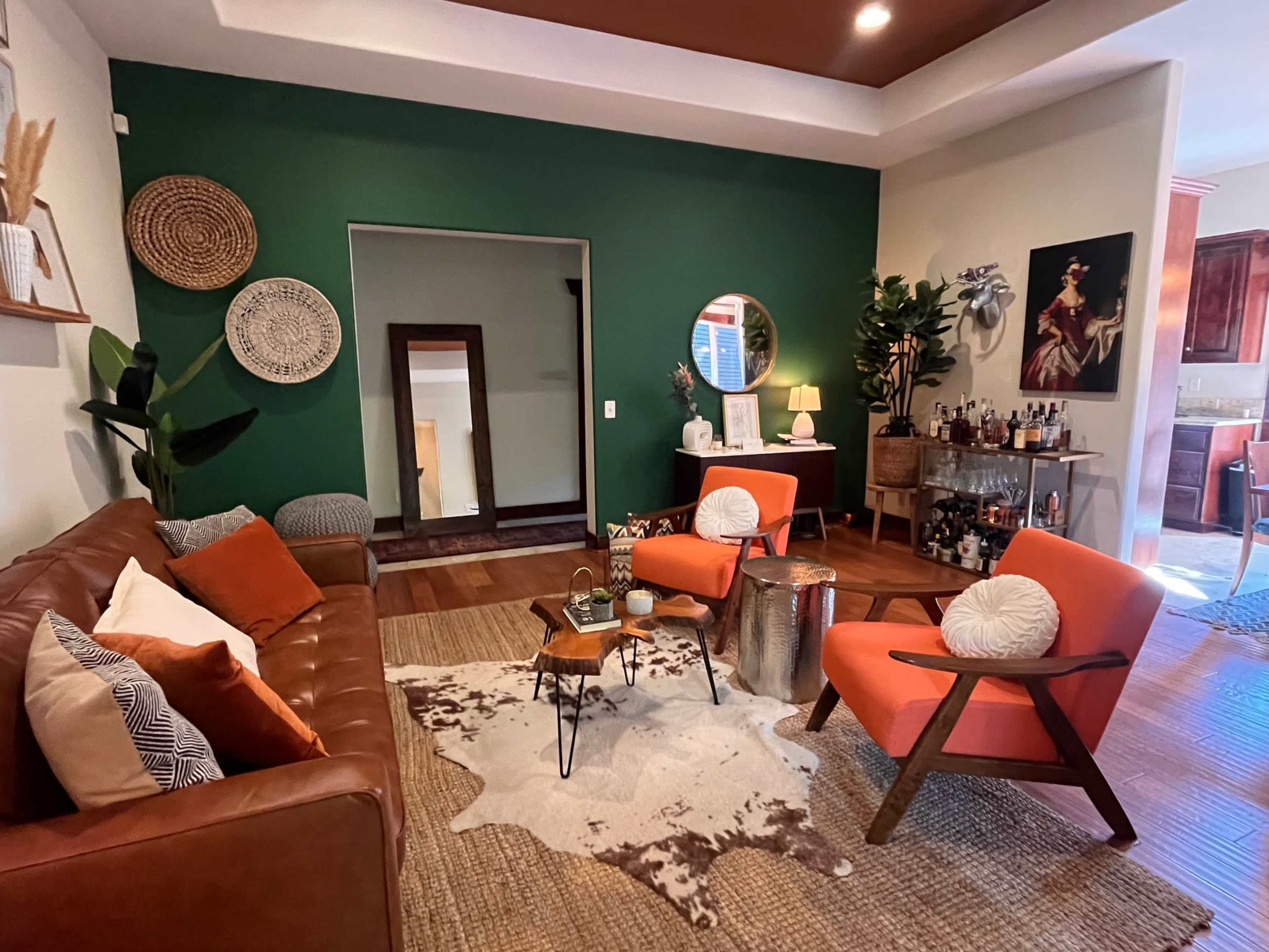 The image shows a modern living room featuring a brown leather sofa, two orange armchairs, a coffee table on a cowhide rug, and decorative wall hangings against a green accent wall.