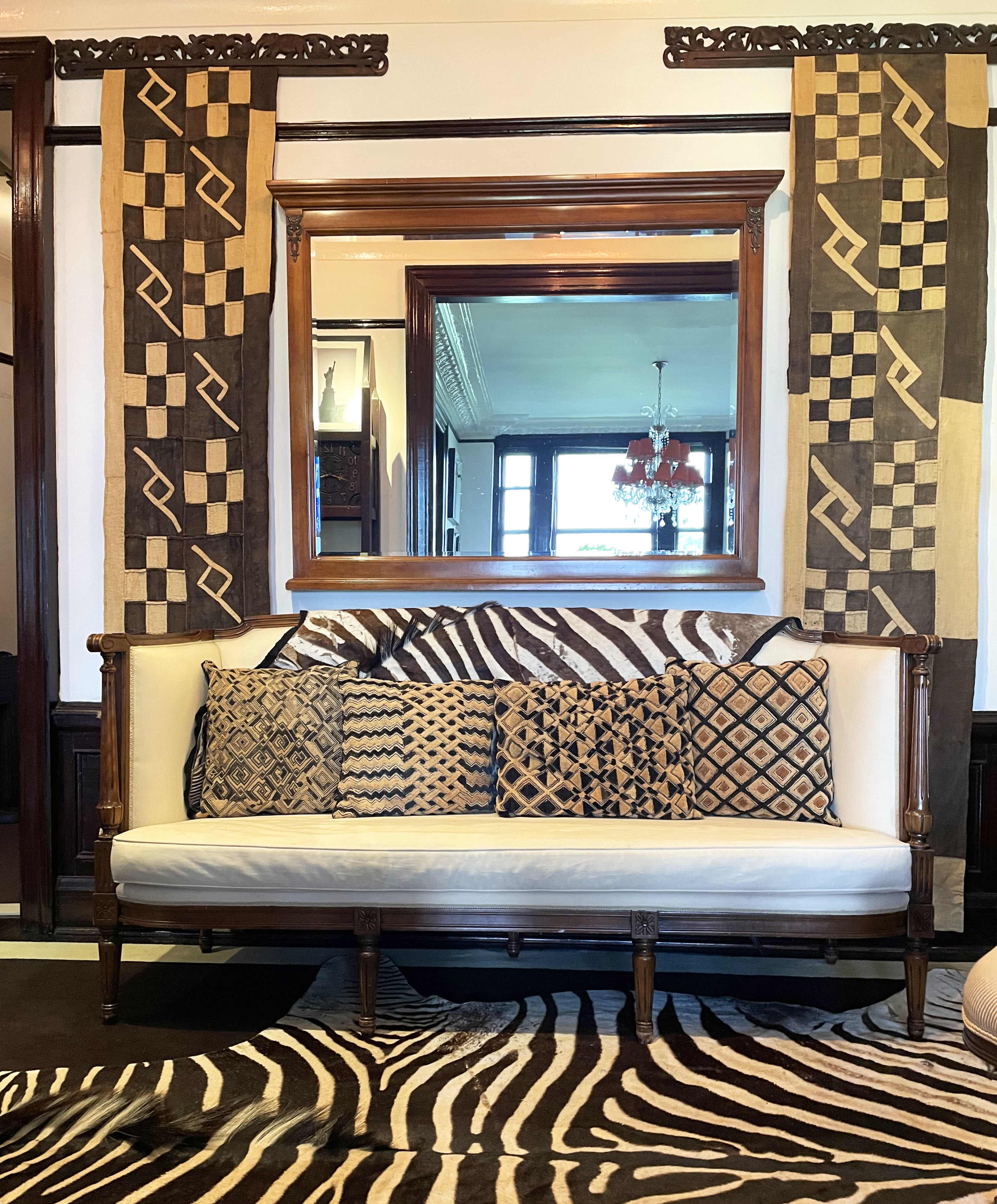 A stylish room featuring a white sofa with patterned cushions in front of a large mirror, flanked by decorative wall hangings and a zebra-print rug.