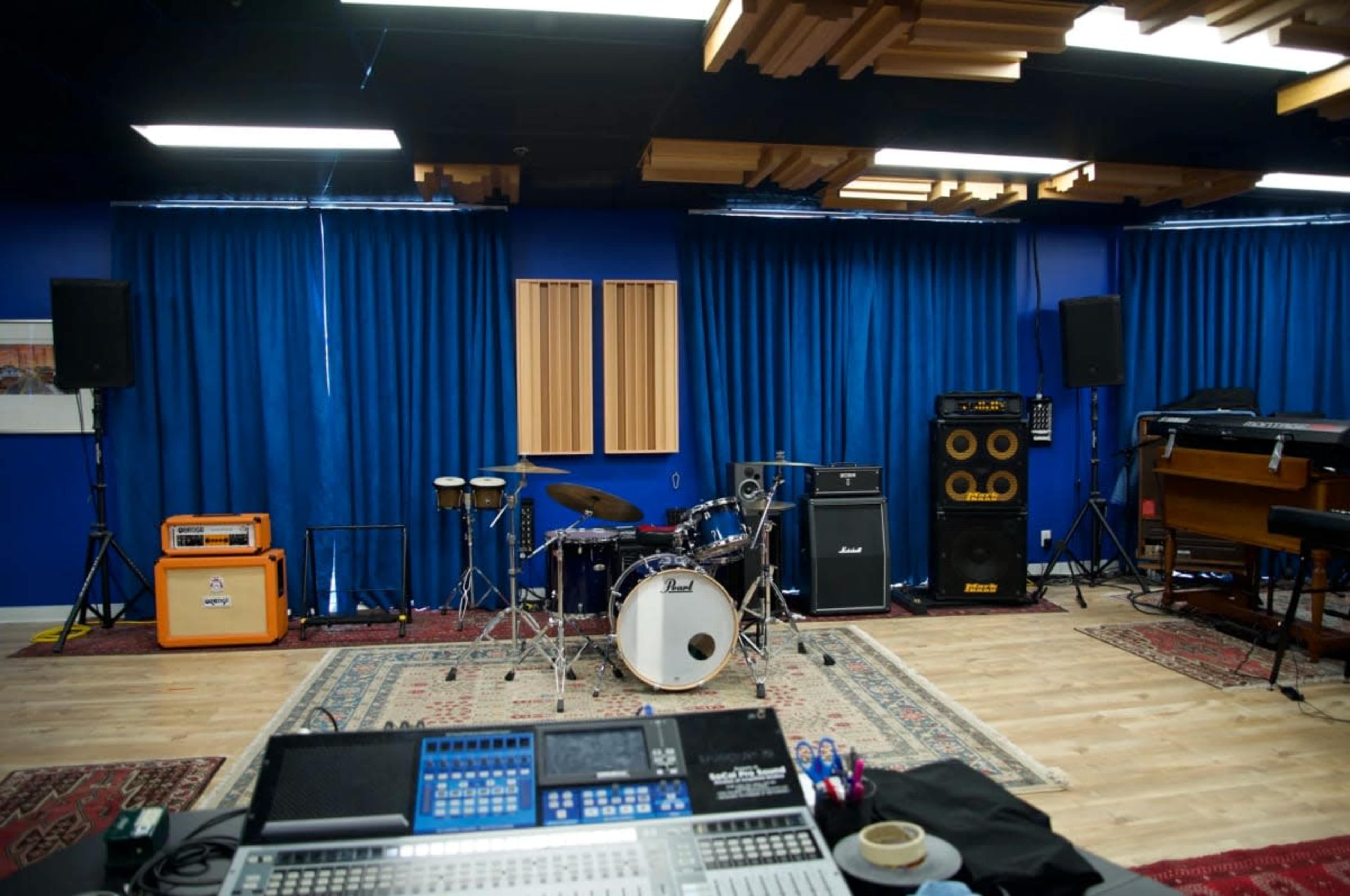 The image shows a music studio with a drum set, various amplifiers, and mixing equipment, arranged in a spacious room with blue walls and wooden ceiling paneling.