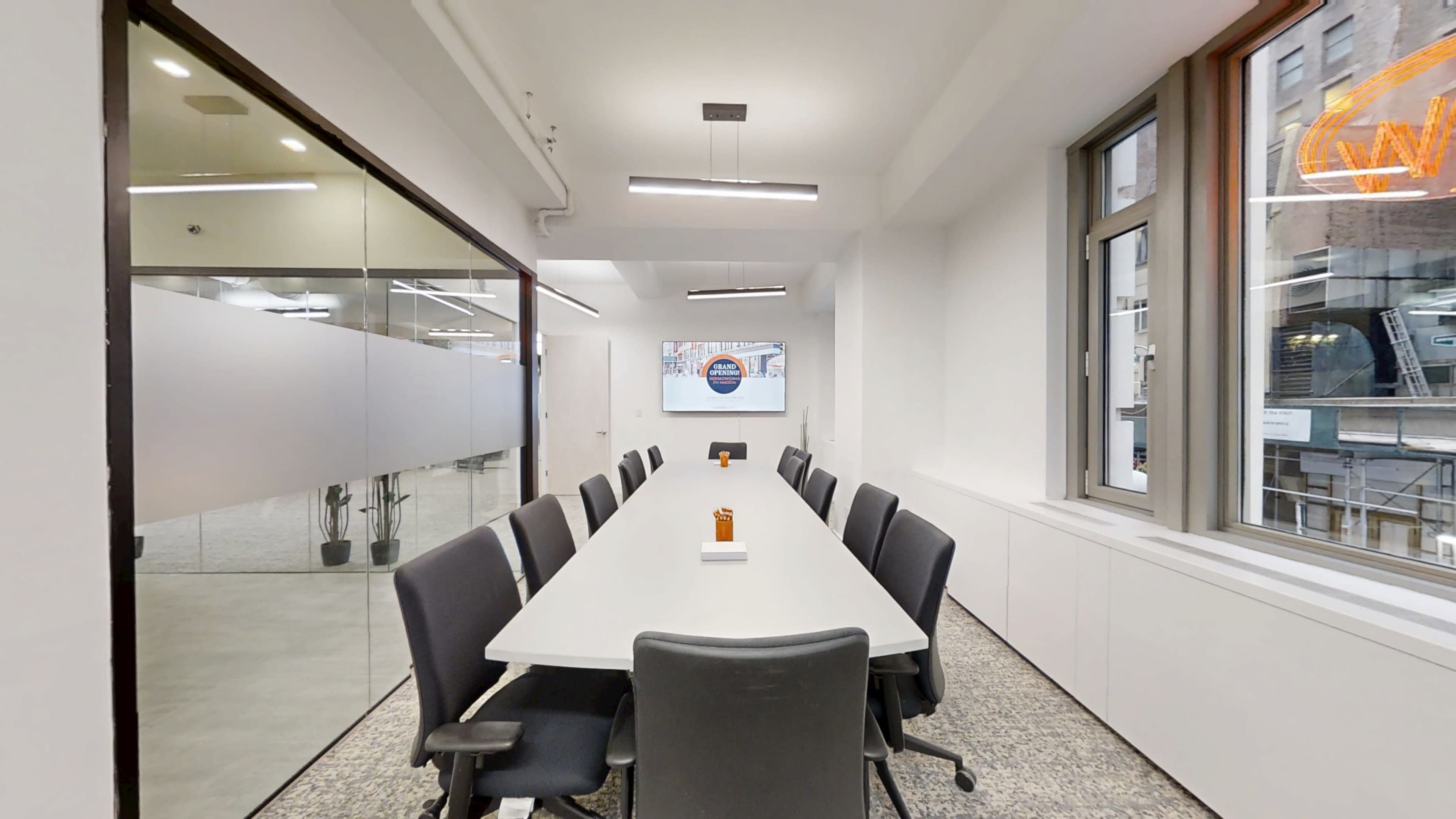 The image shows a modern conference room with a long table and several black chairs, featuring glass walls and large windows.
