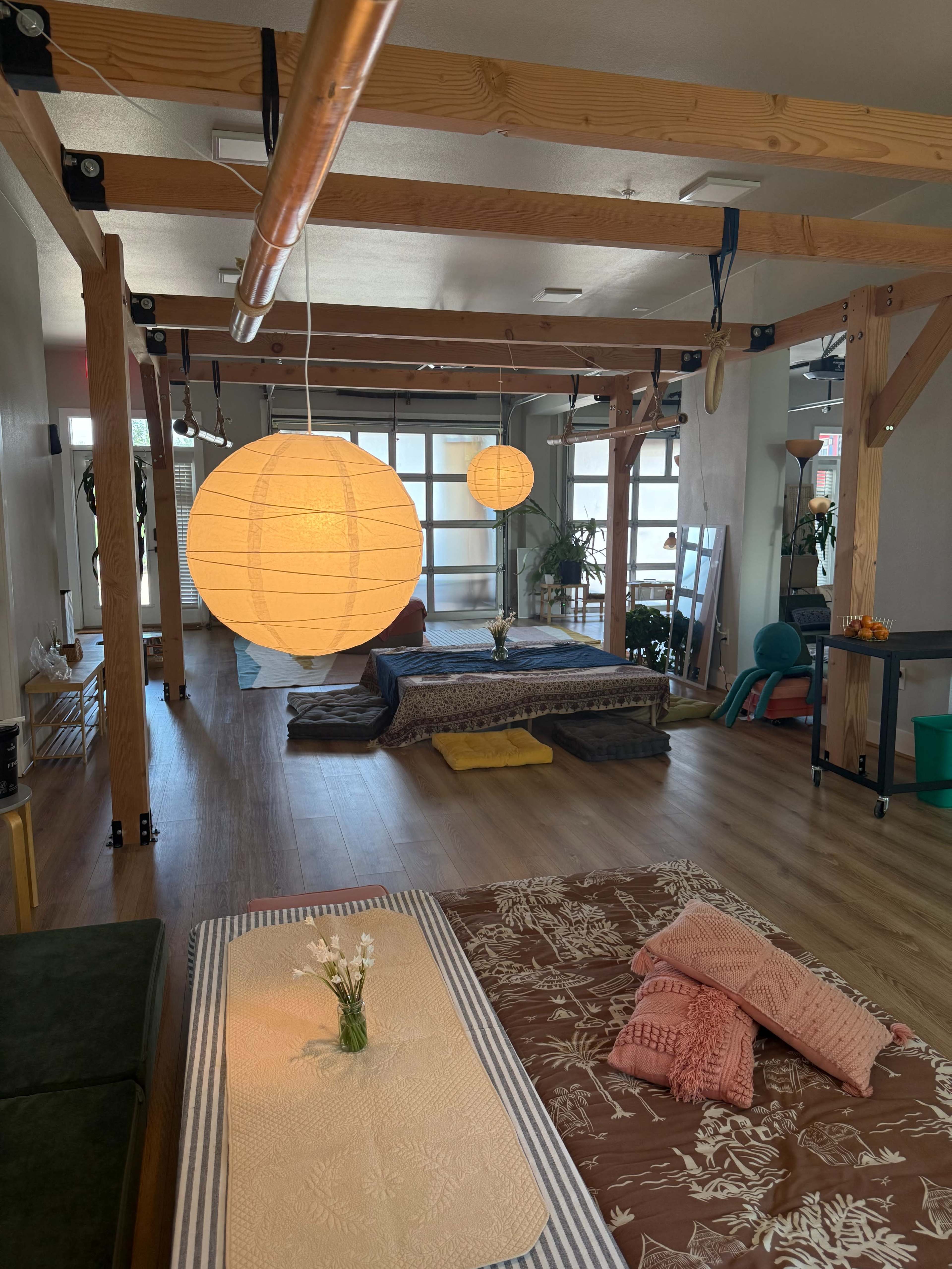 The image shows a spacious room with wooden beams, soft floor mats, and hanging paper lanterns, creating a cozy atmosphere.