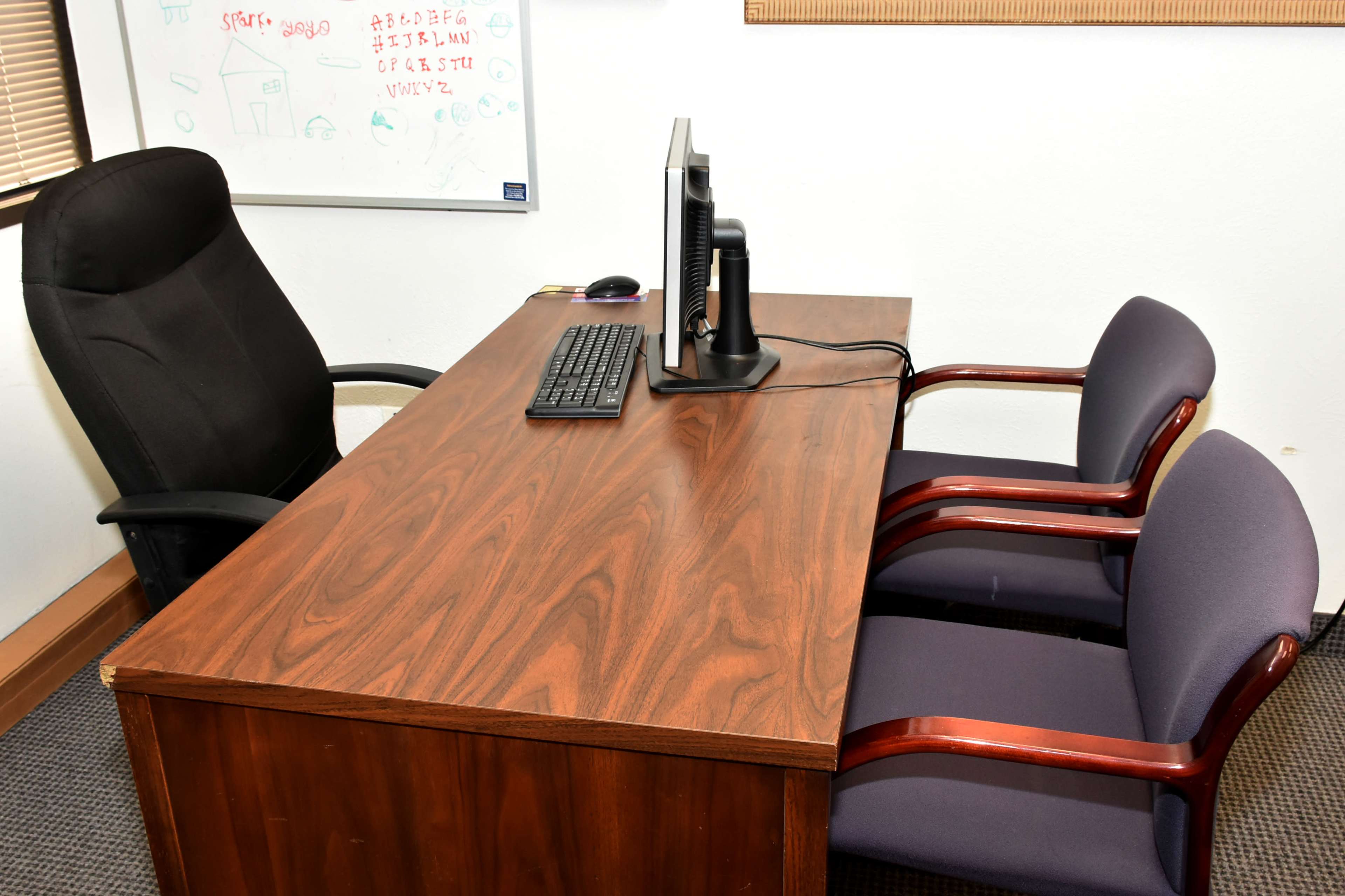 A wooden desk with a computer monitor and keyboard is positioned in front of an office chair and two purple guest chairs.