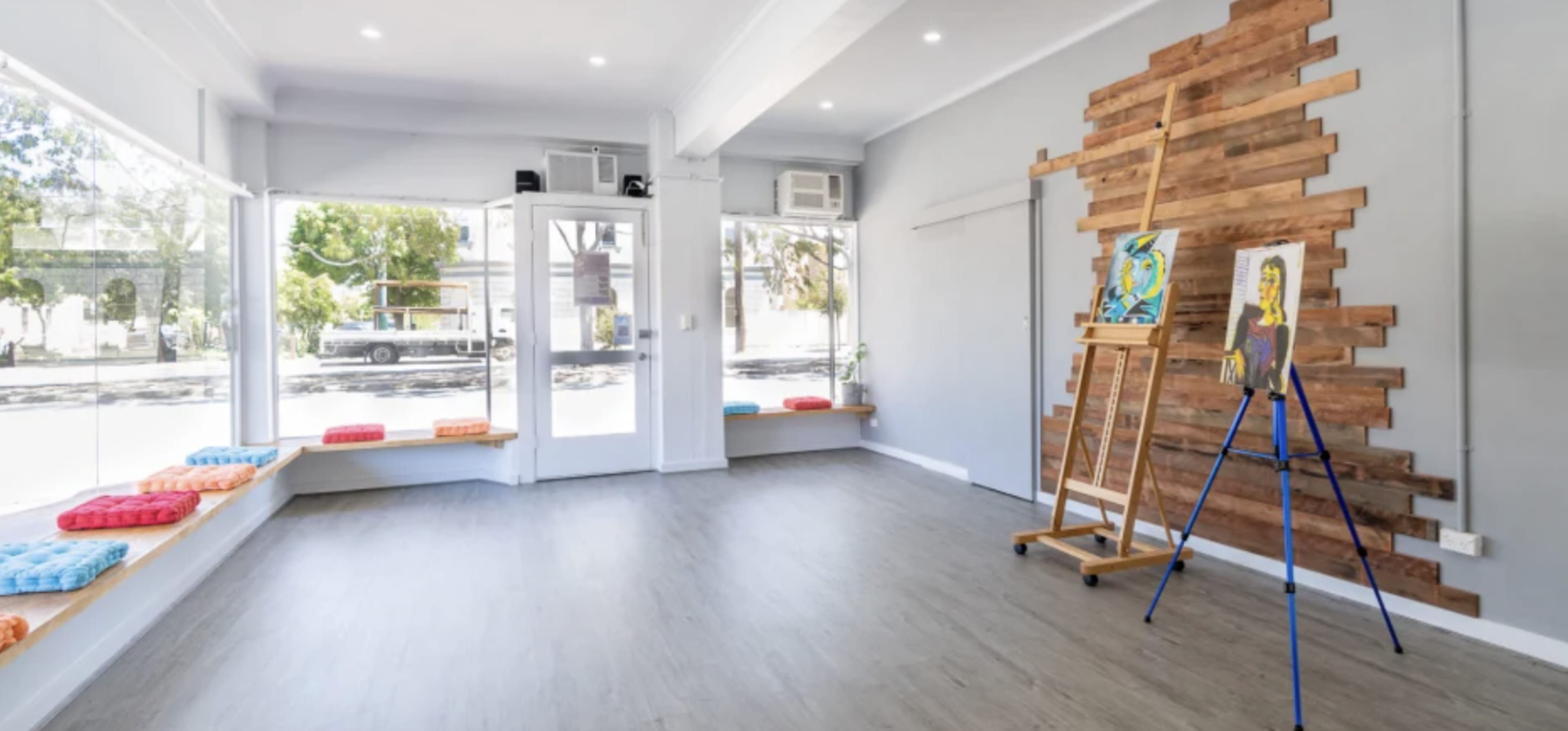 The image shows a spacious, minimally furnished room with large windows, an easel displaying artwork, and wooden accents on the wall.