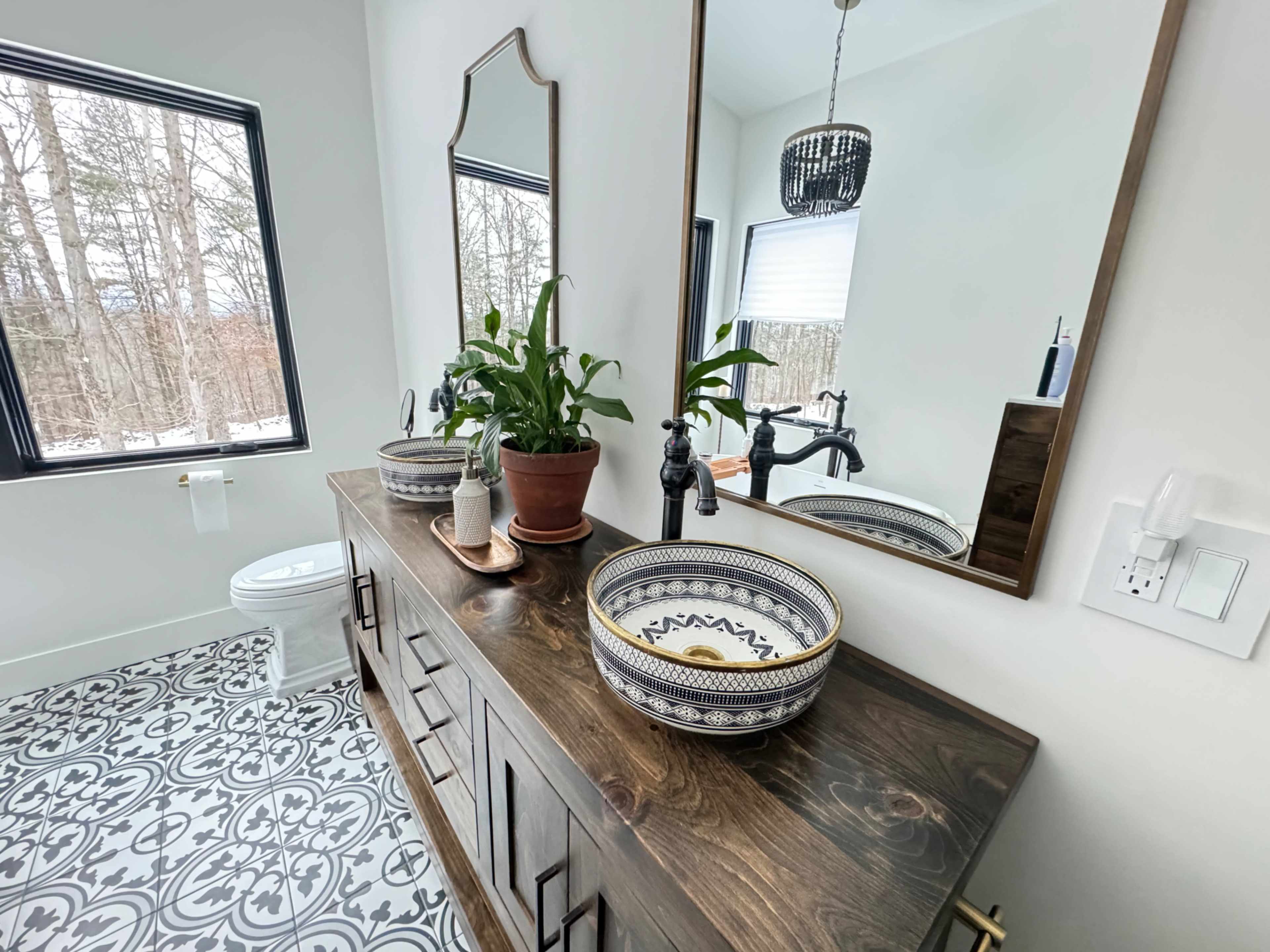 The image shows a modern bathroom featuring a wooden vanity with a decorative sink, two mirrors, a potted plant, and patterned tile flooring.