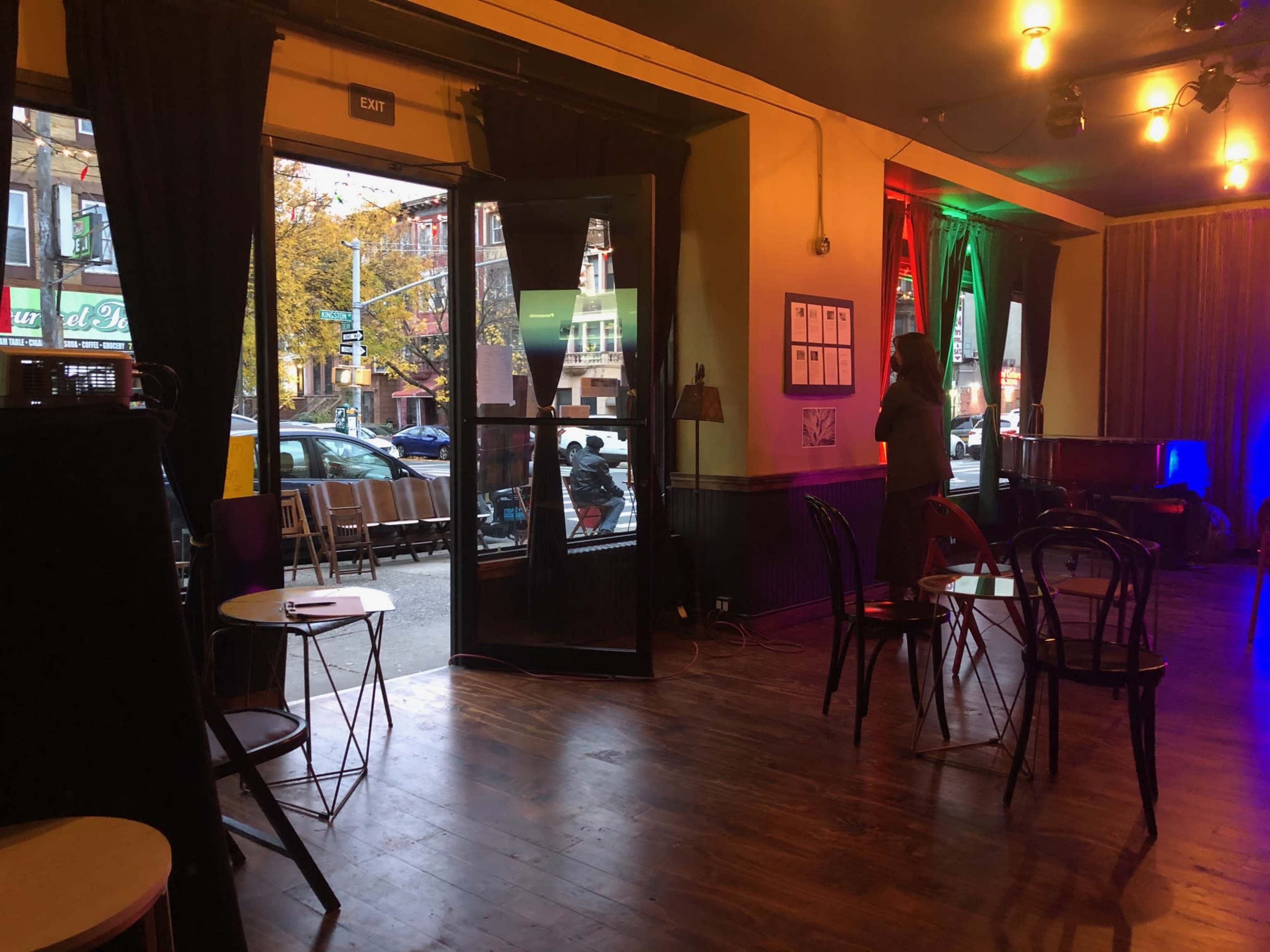 A person stands near an open door in a dimly lit room with wooden floors and chairs arranged around small tables.