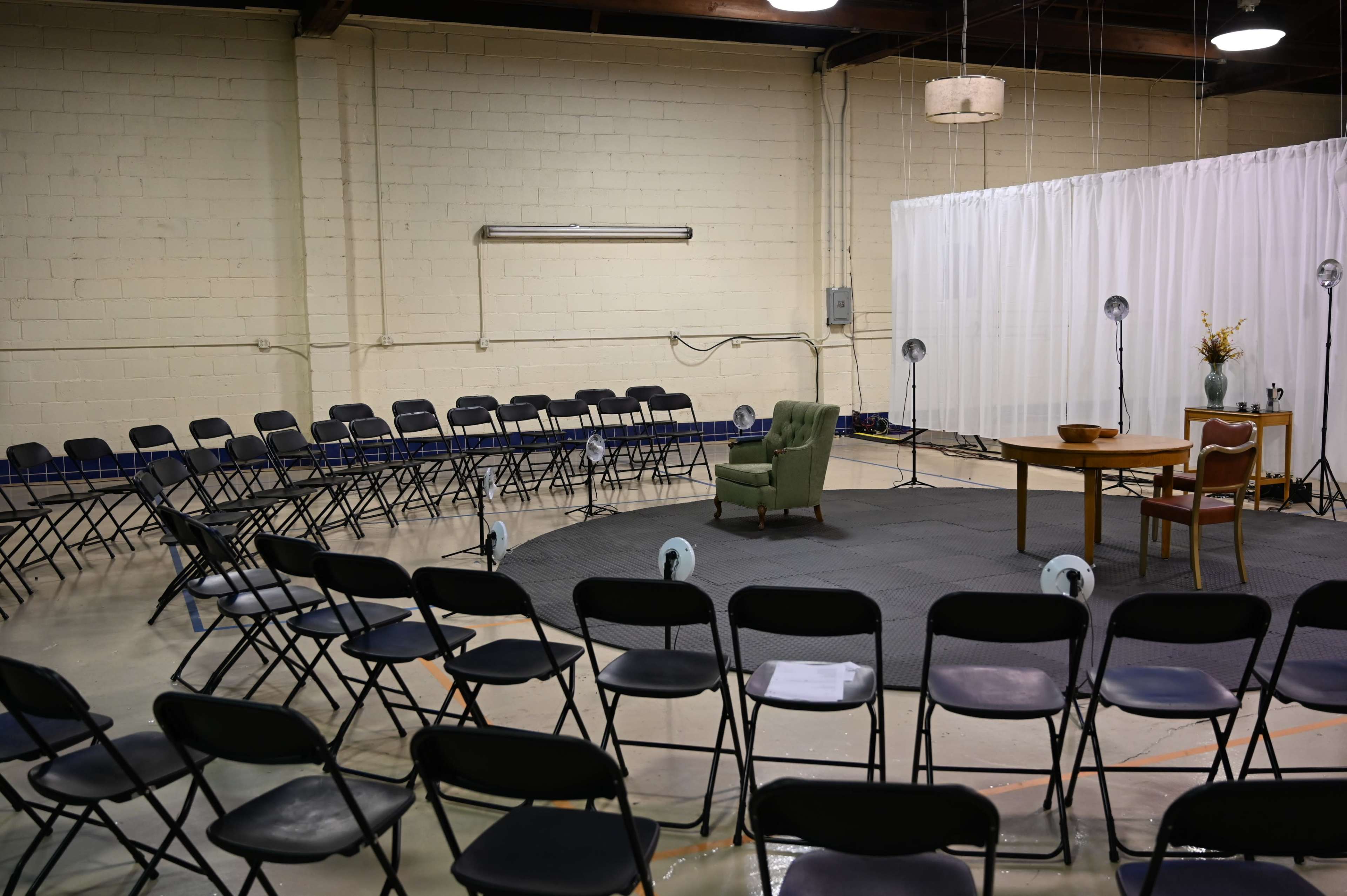 The image shows a circular arrangement of folding chairs surrounding a central area with a green armchair and a table, all set within a large, well-lit room.