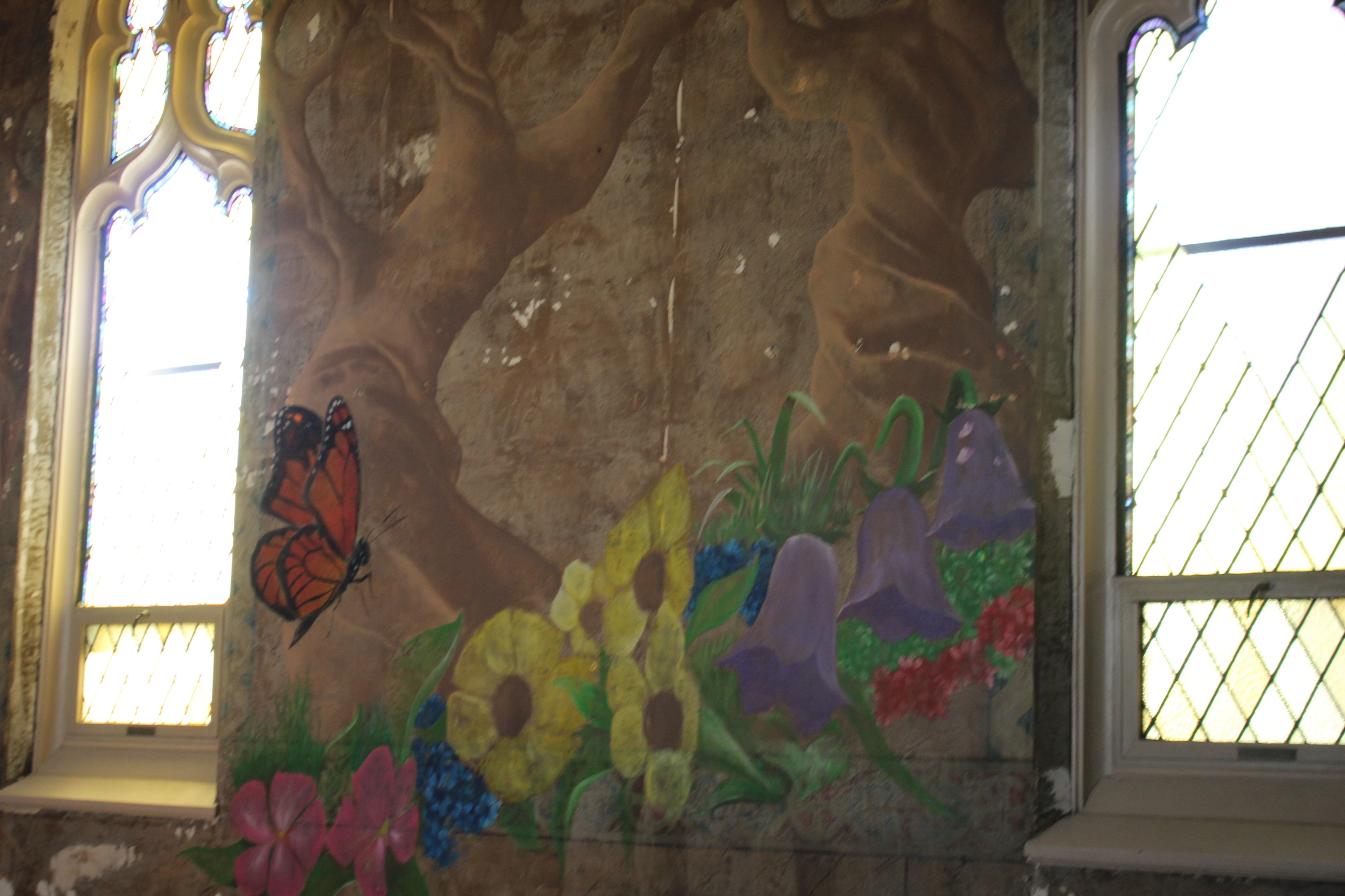 The image shows a mural featuring a butterfly and various flowers painted on a wall with large windows in the background.