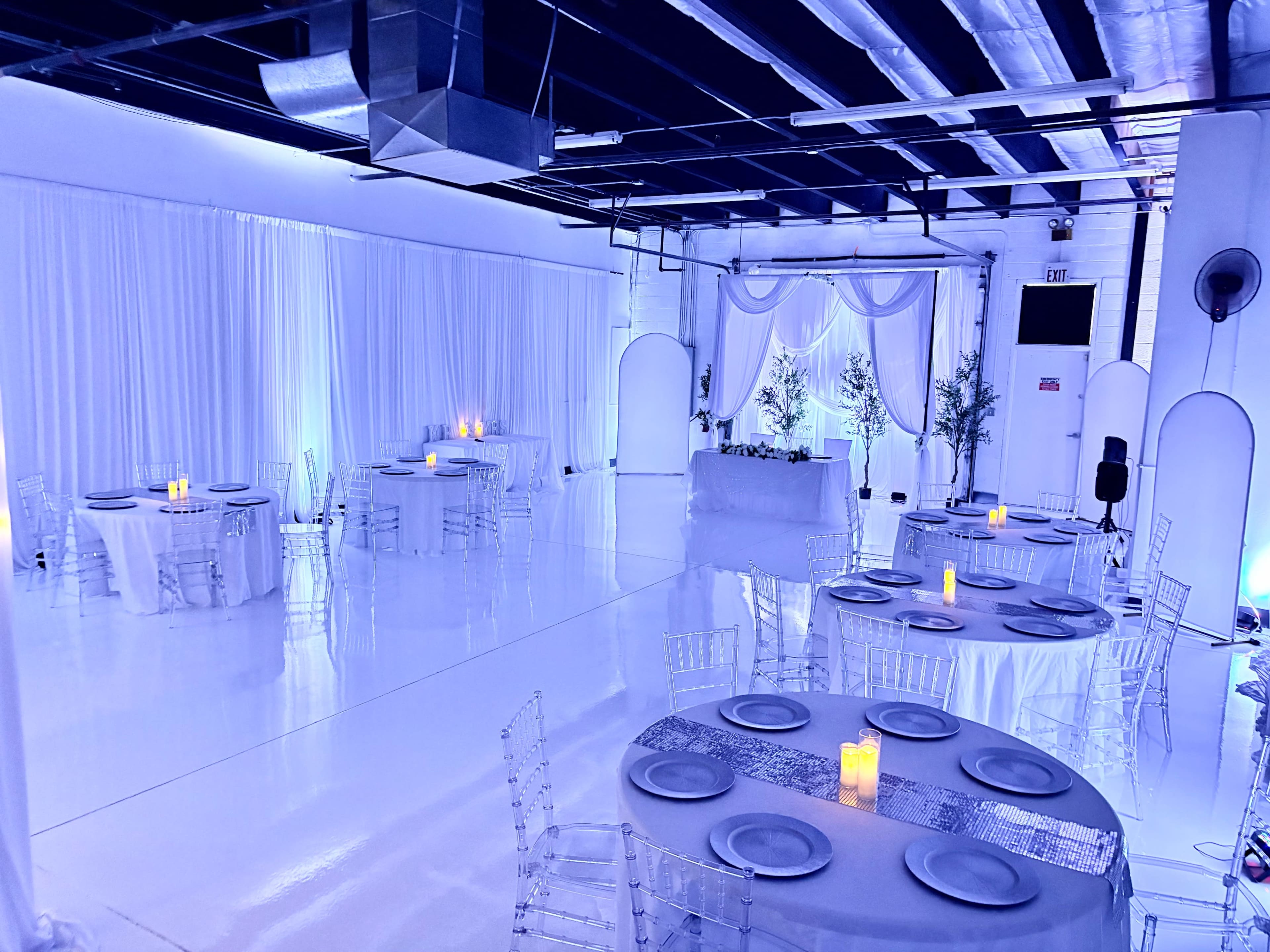 The image shows a well-lit event space with white walls, round tables set with candles, and elegant decor, including sheer curtains and greenery.