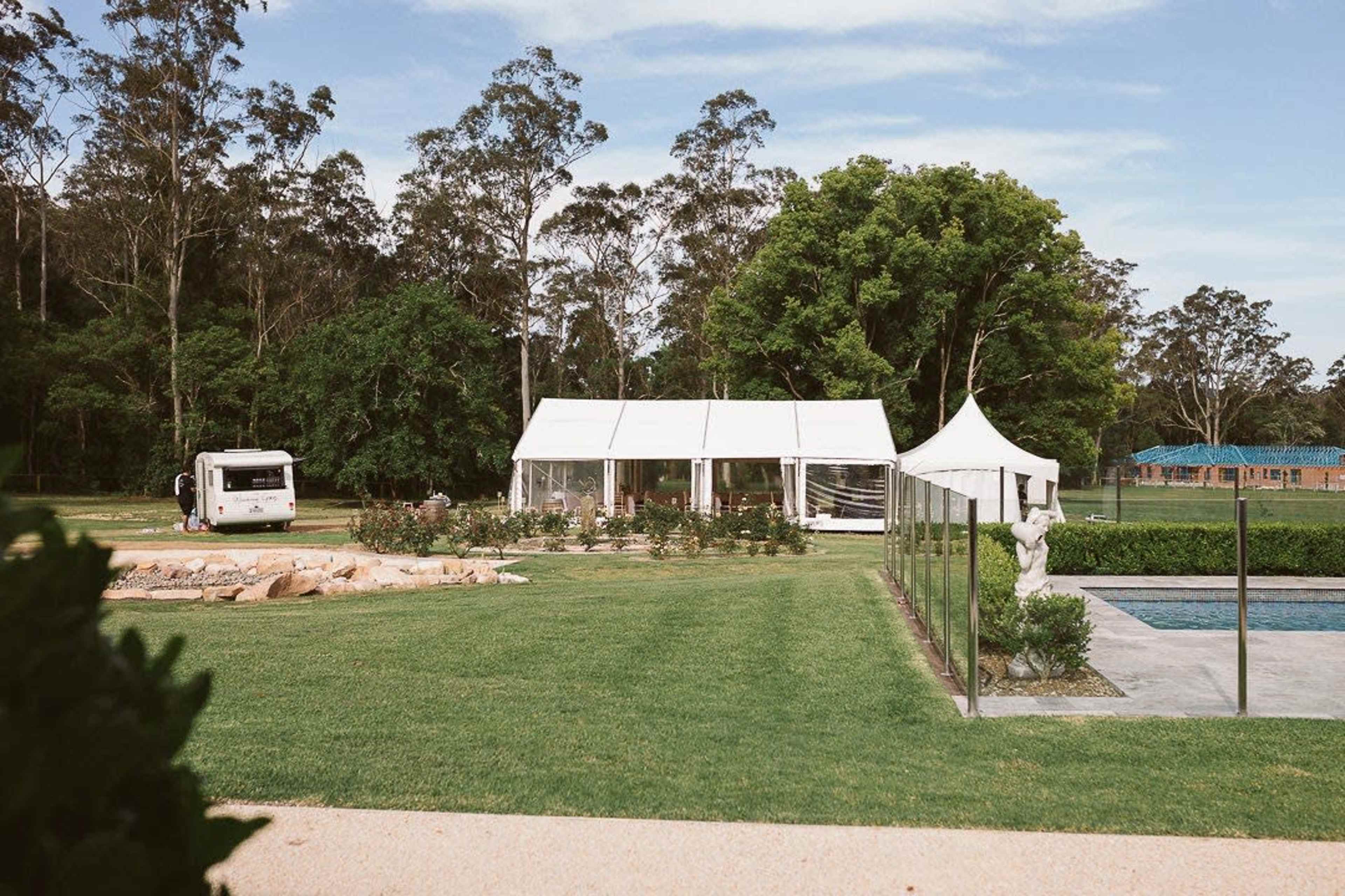 A white tent and a smaller canopy are set up near a pool, surrounded by a well-maintained lawn and trees in the background.