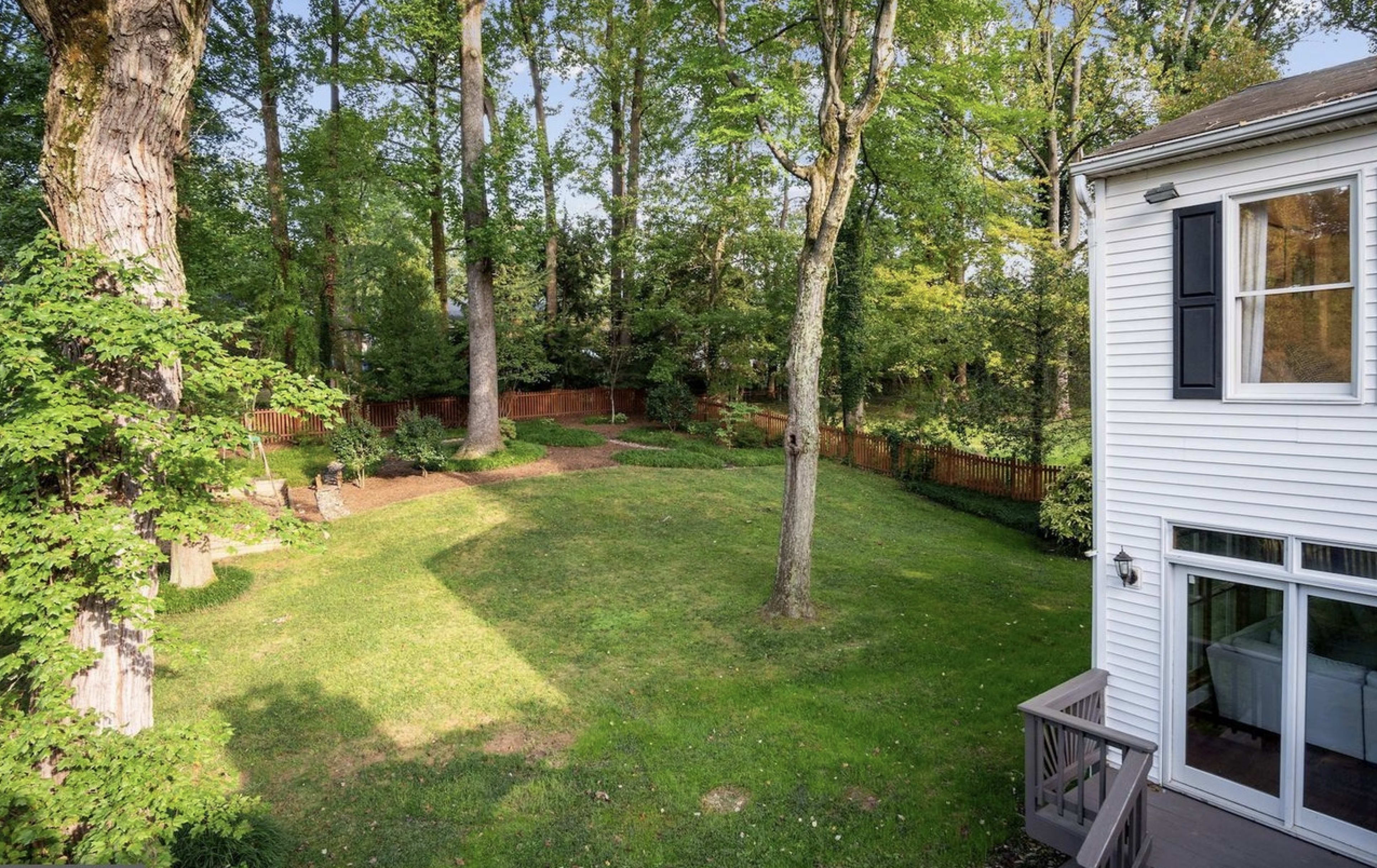 The image shows a spacious backyard with green grass, a few trees, and a wooden fence.