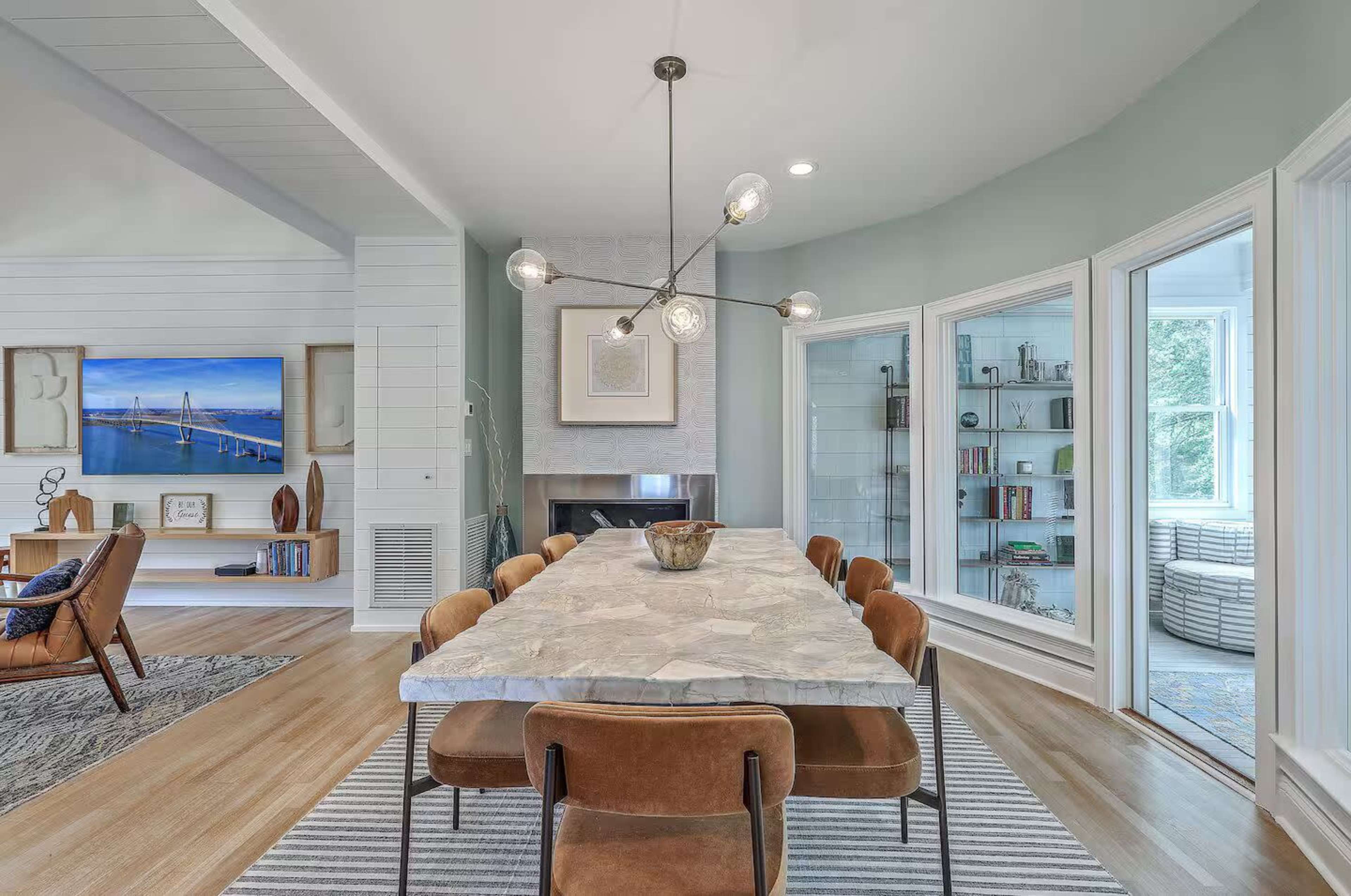 The dining area features a large stone table surrounded by brown chairs, illuminated by an artistic light fixture, with large windows and a modern aesthetic.