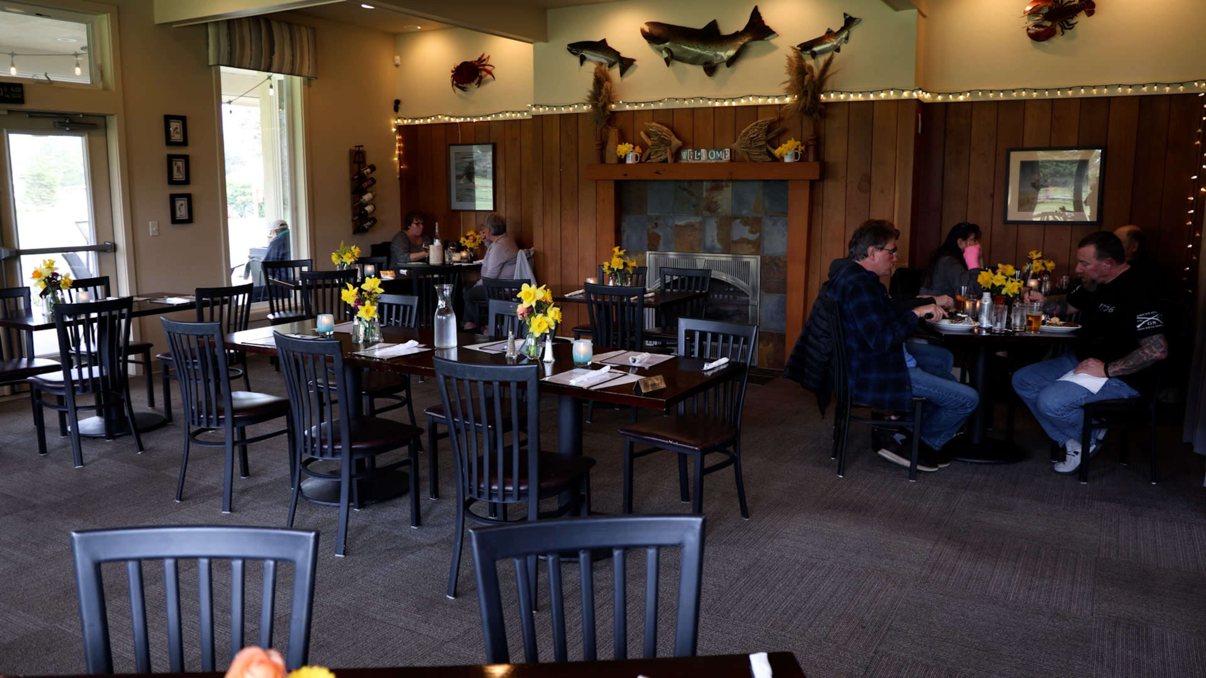 The image shows a restaurant interior with wooden walls, tables set for dining, and two groups of people eating at separate tables, surrounded by decorative fishing-themed art.