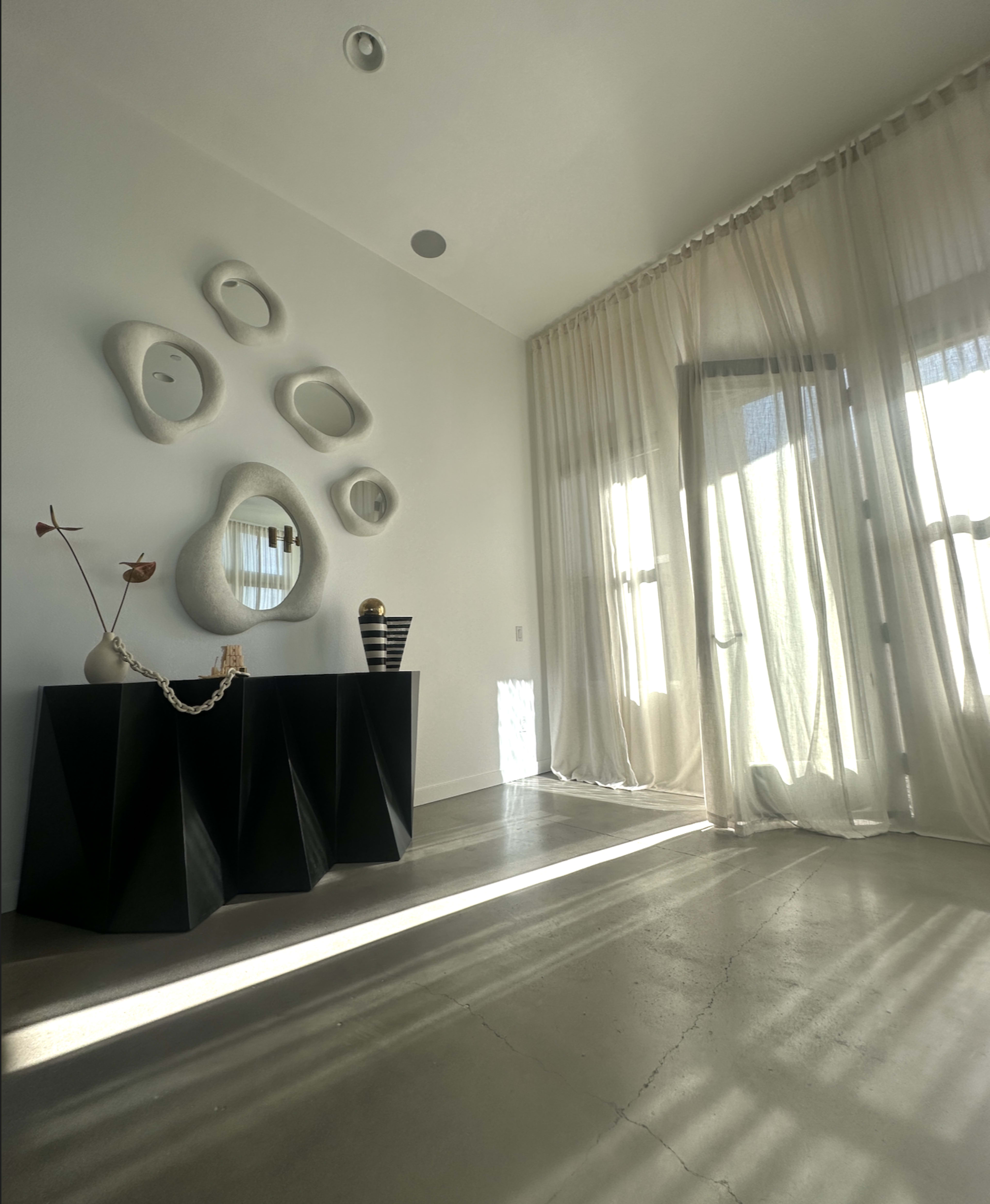 The image shows a minimalist interior with a dark console table beneath a wall adorned with textured, irregular mirrors and sheer curtains partially covering large windows.