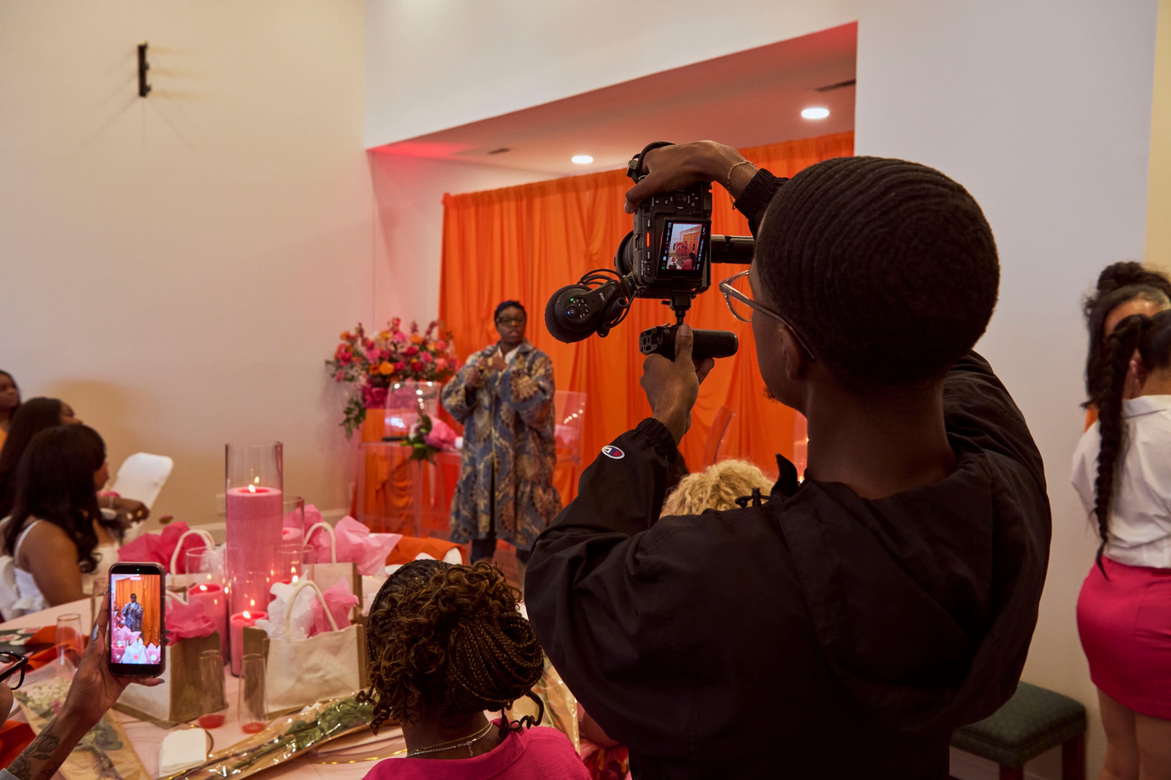 A man holds a camera to capture a speaker at a decorated event with an orange backdrop and tables adorned with gifts.