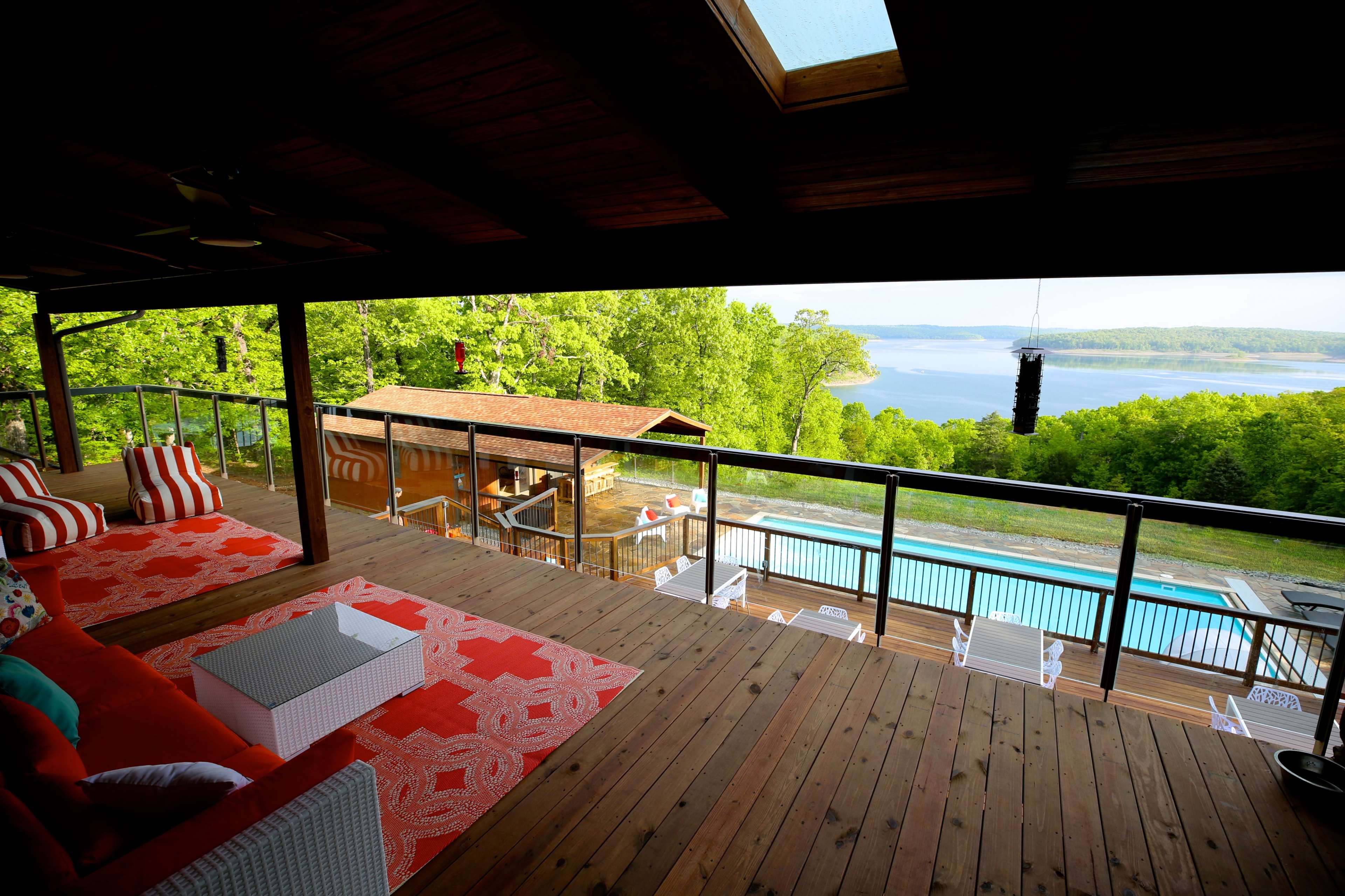The image shows a wooden deck with outdoor seating overlooking a pool and a lake surrounded by greenery.