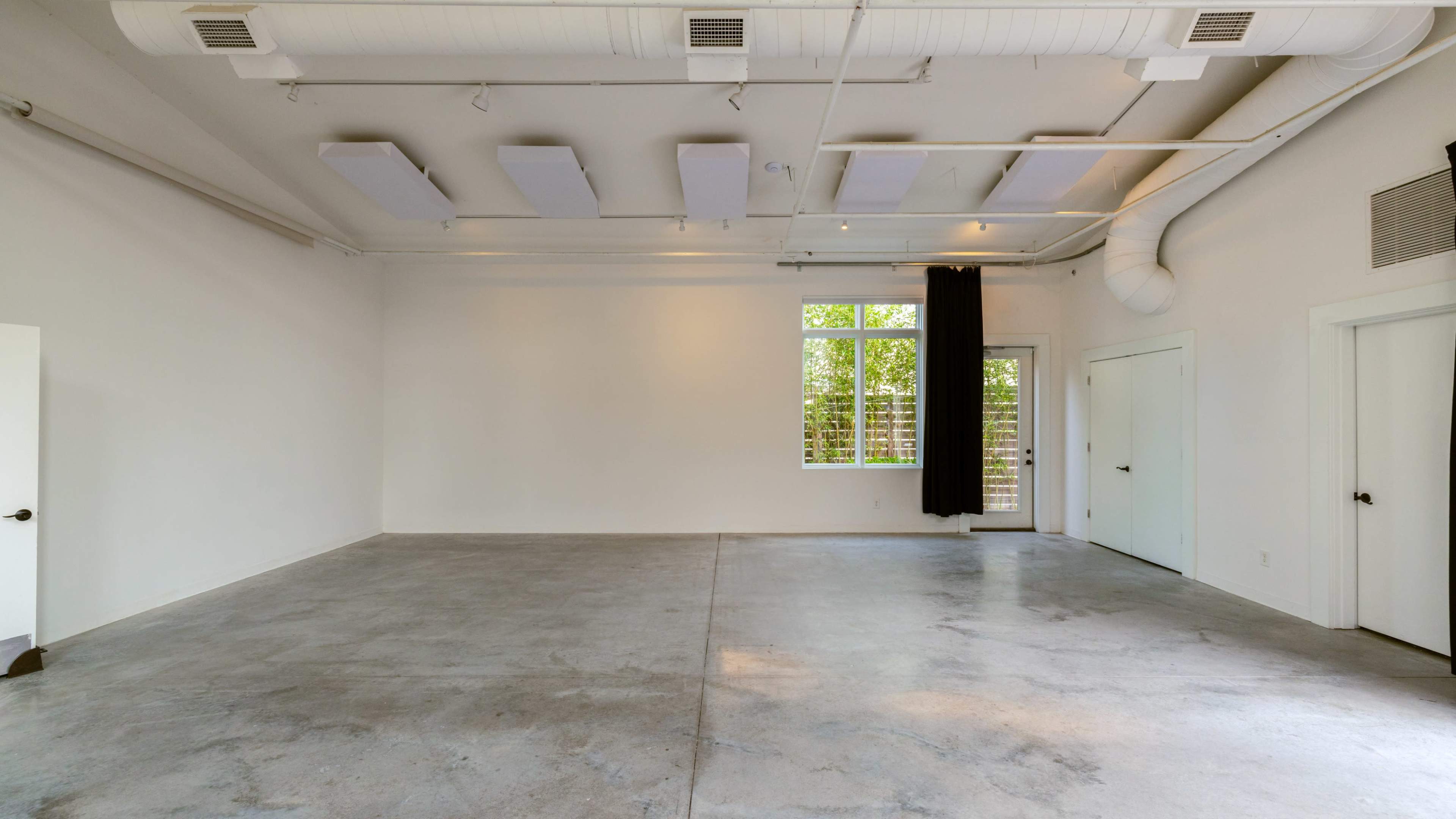 The image shows an empty room with concrete flooring, white walls, and large windows with black curtains.