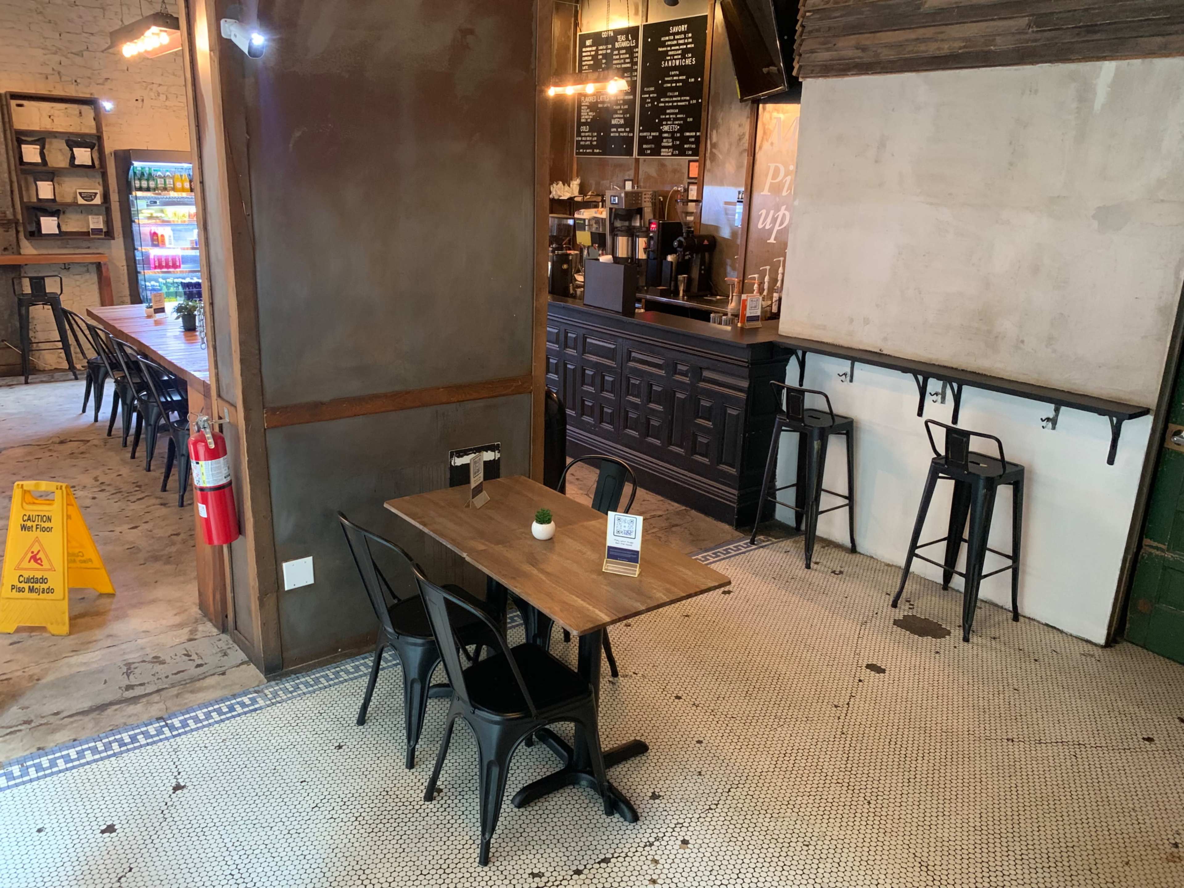 A café interior with a small table and chairs in the foreground, a counter with drinks behind it, and a long communal table in the background.