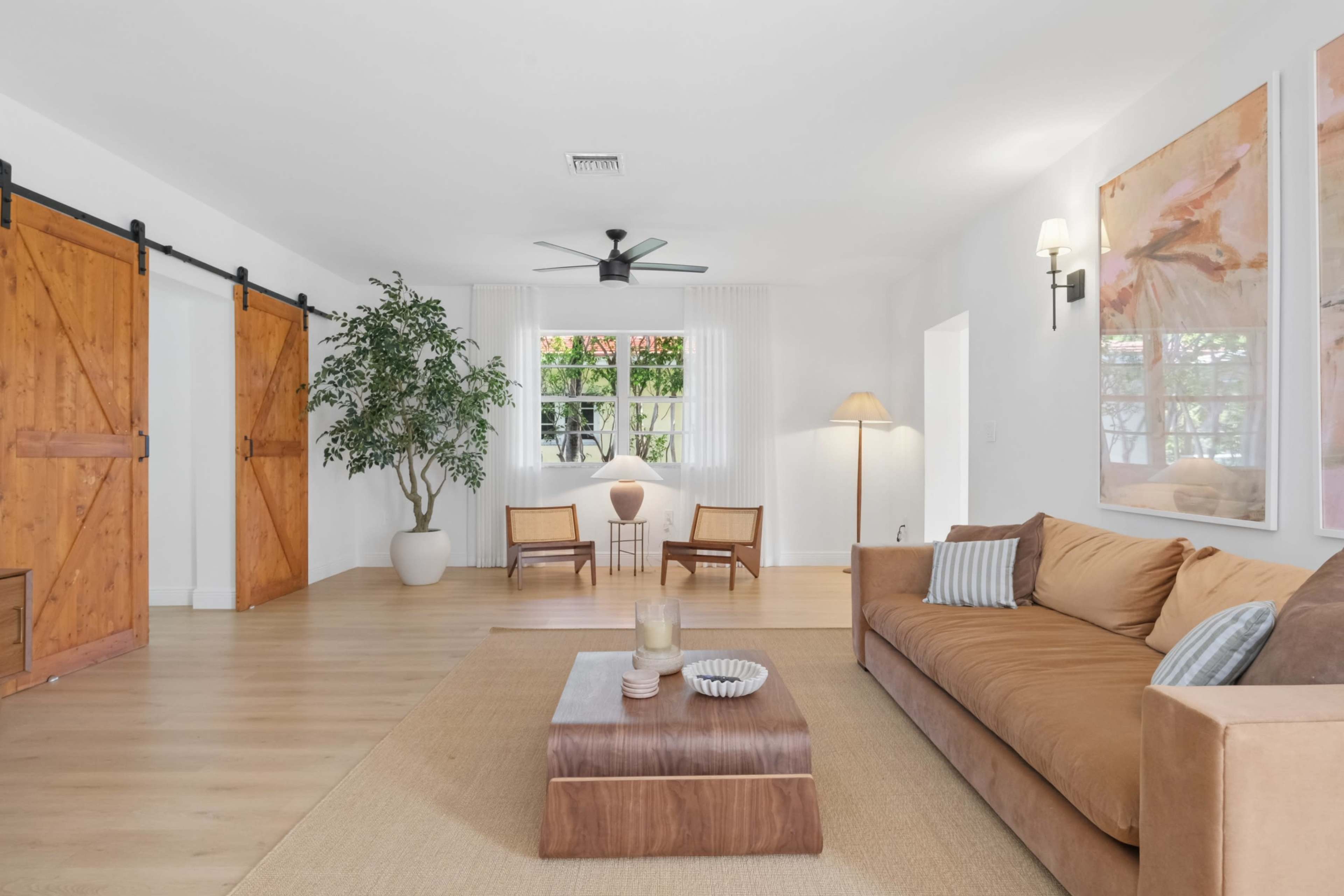 A modern living room features a brown sofa, a wooden coffee table, plants, two armchairs, and sliding barn doors.