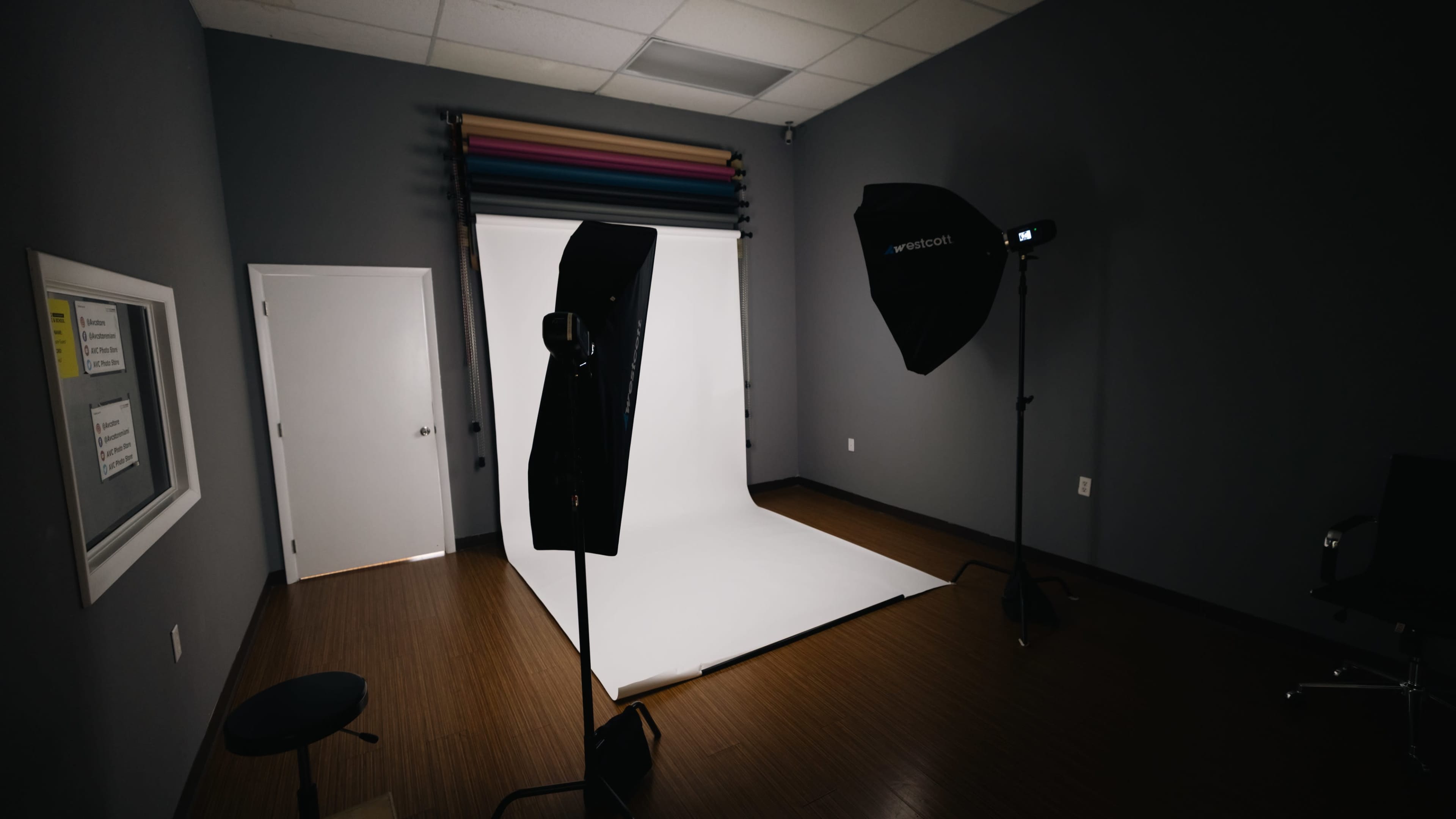 The image shows a photography studio with a white backdrop, two lighting stands, and a black chair against a gray wall.