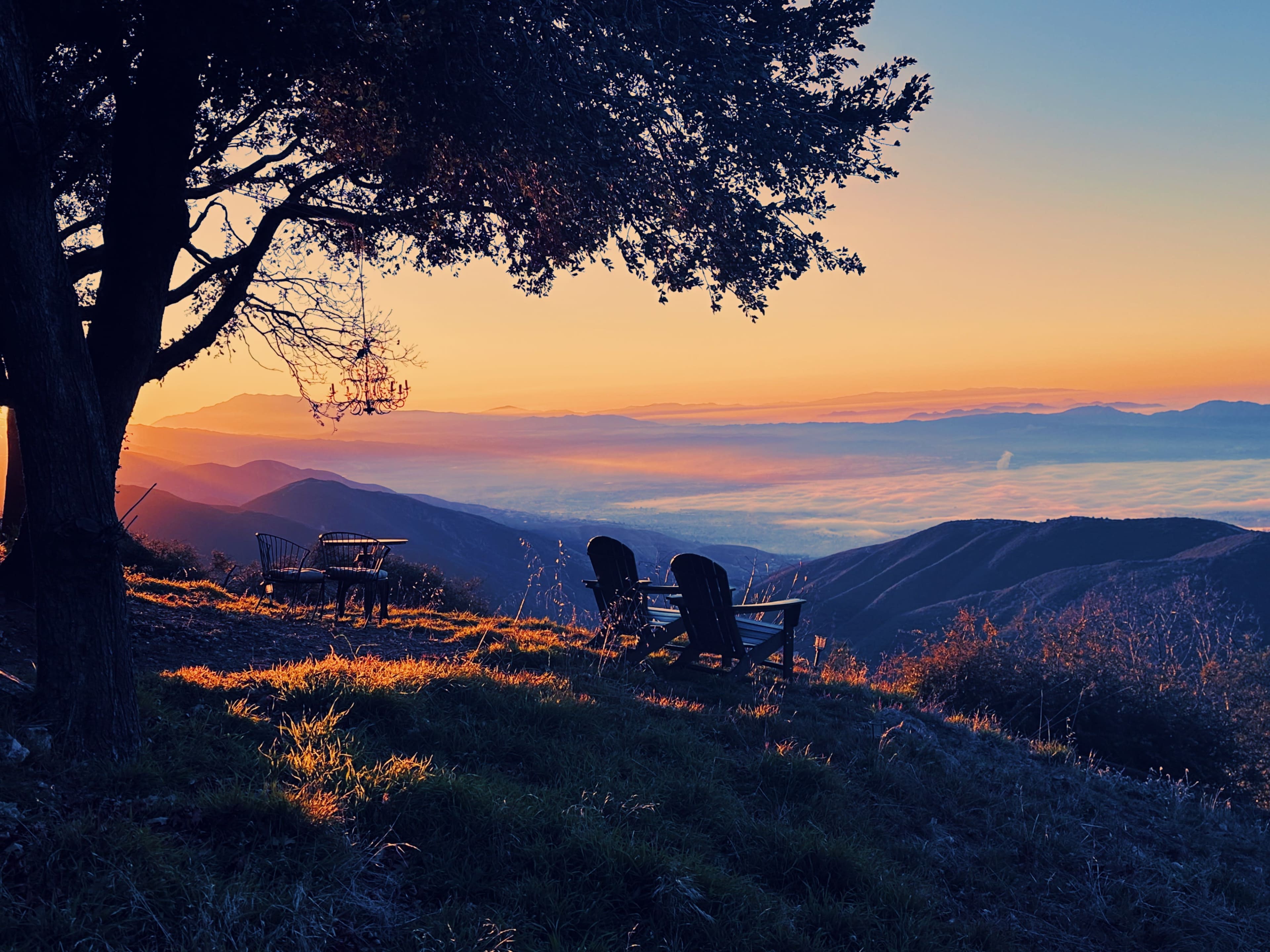 Two wooden chairs sit beside a tree overlooking a valley at sunset, with mist rising from the landscape below.