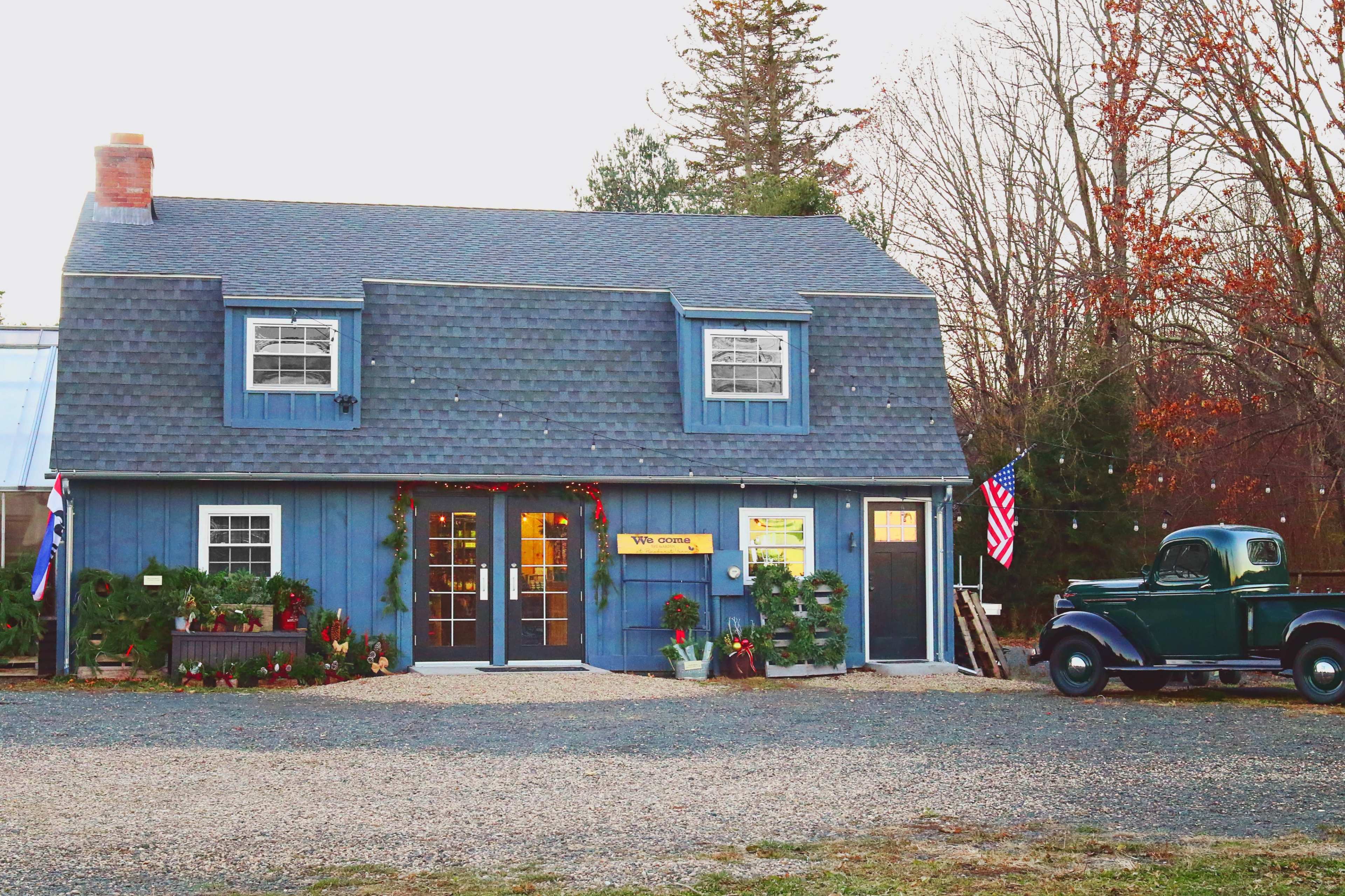 A blue two-story building with a sloped roof displays holiday decorations, while an old green truck is parked nearby on a gravel driveway.